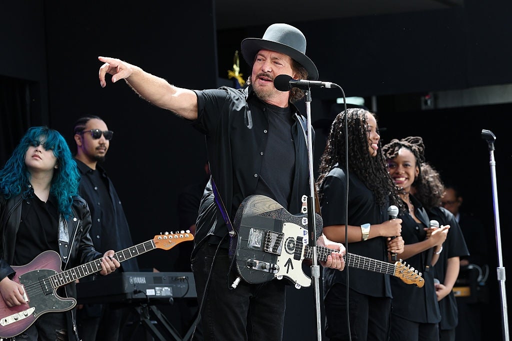 Musiker som opptrer på scenen med gitar, iført en bredbremmet hatt og helsvart antrekk; backup sangere og band akkompagnerer ham