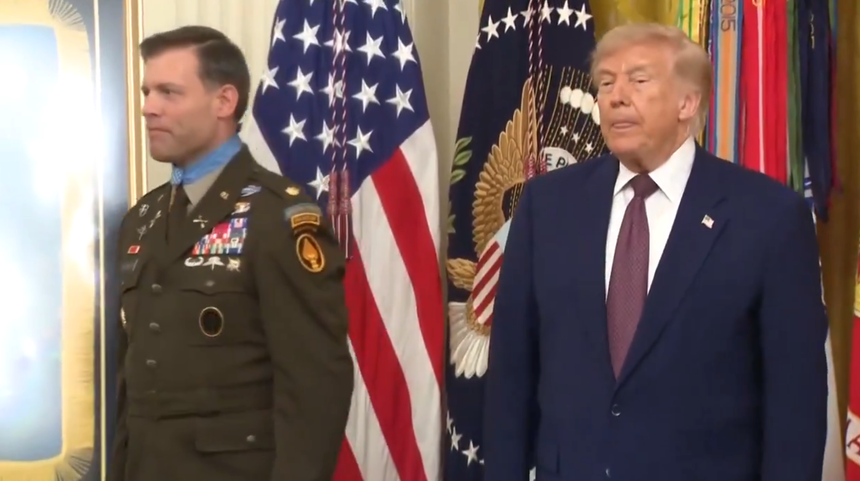 To menn står i en formell setting, med den ene i militæruniform og den andre i dress. USA-flagg er synlige i bakgrunnen