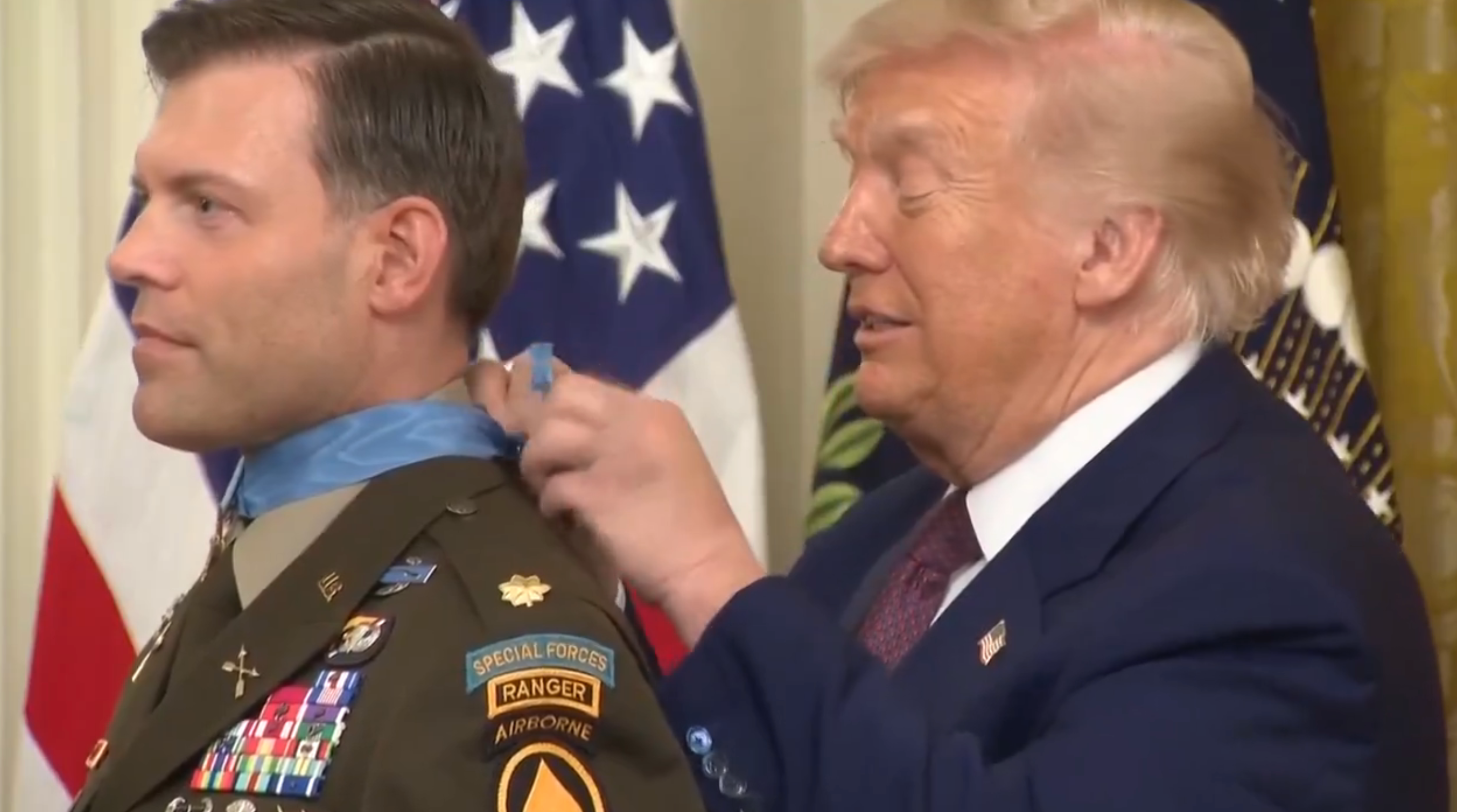 En mann i militæruniform mottar en medalje fra en annen mann i dress under en formell seremoni. Flagg er synlige i bakgrunnen
