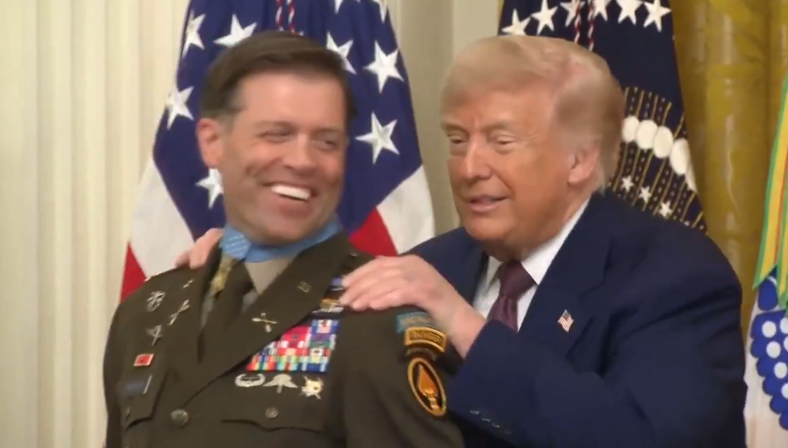 To personer smilende, den ene i militærdrakt med medaljer, den andre i dress. De ser ut til å være på et formelt arrangement med flagg i bakgrunnen