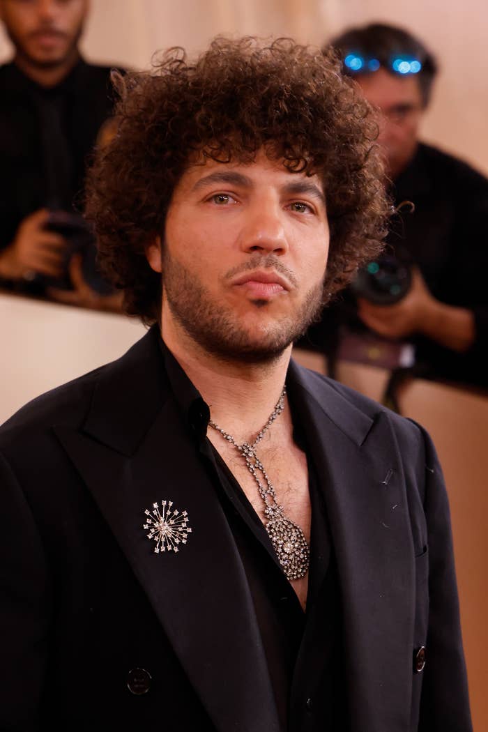 Benny Blanco with curly hair wears a stylish suit, ornate necklaces, and a decorative brooch, posing on the red carpet