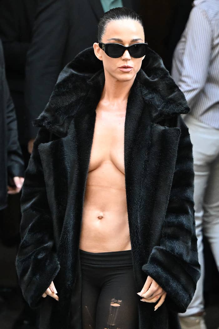 Person wearing a long fur coat with no shirt underneath, stylish sunglasses, and form-fitting pants in a public setting