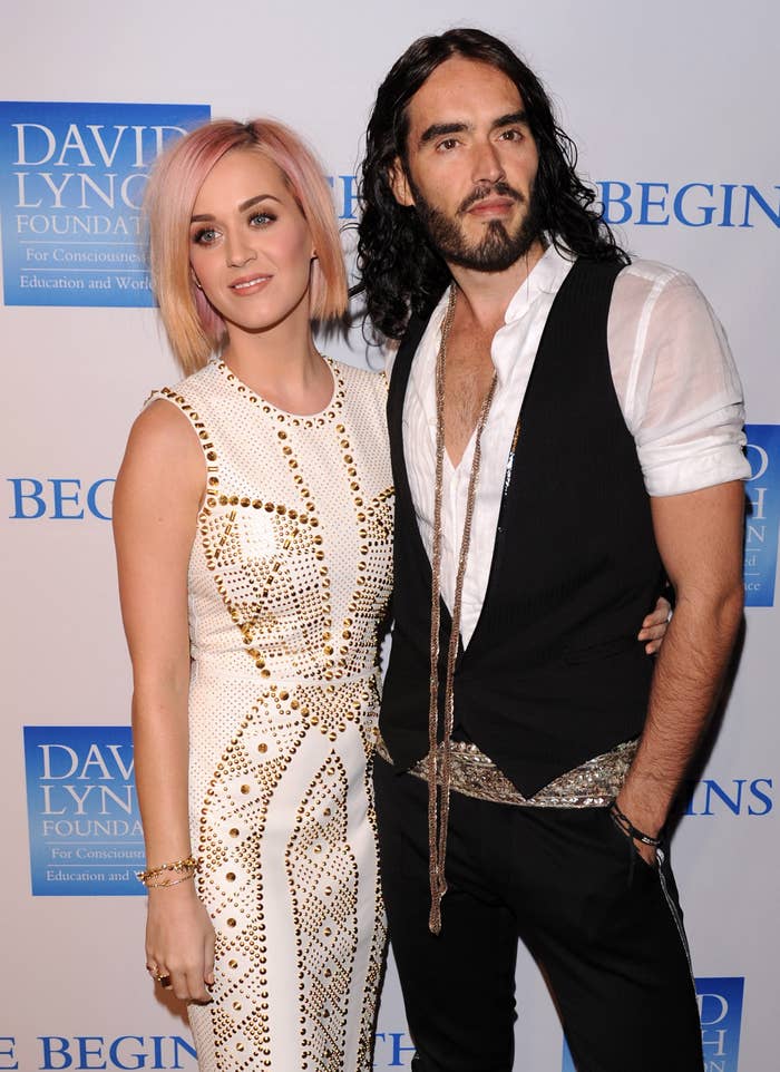 Two people on a red carpet; one wears a studded dress, the other a vest and scarf. They pose in front of a backdrop with text