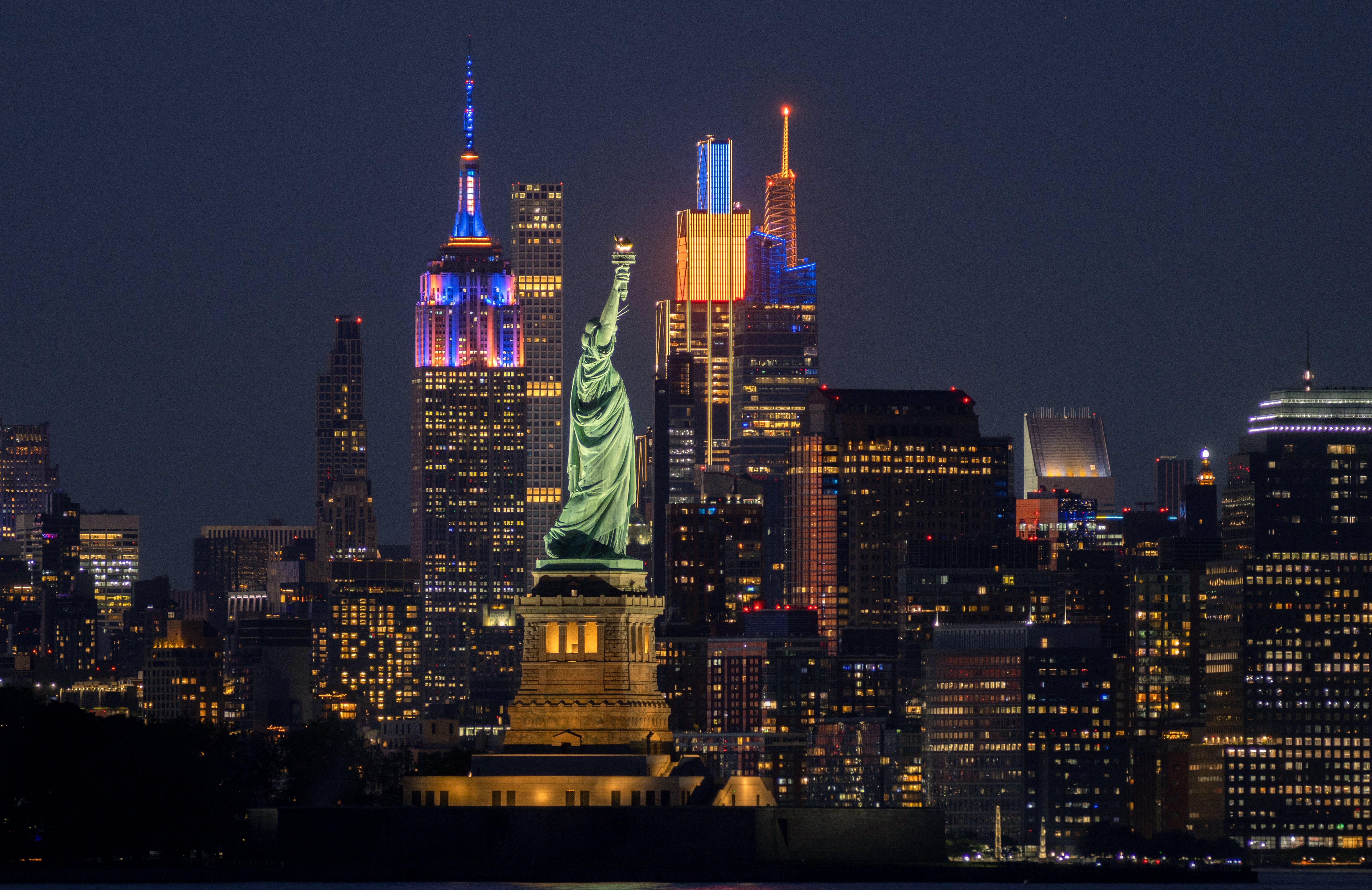 Frihetsgudinnen om natten med opplyste skyskrapere i NYC i bakgrunnen