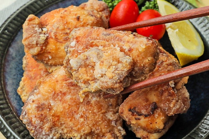 黒い皿に盛られた唐揚げ。箸で一つが持ち上げられており、背景にはトマトとレモンが添えられています。