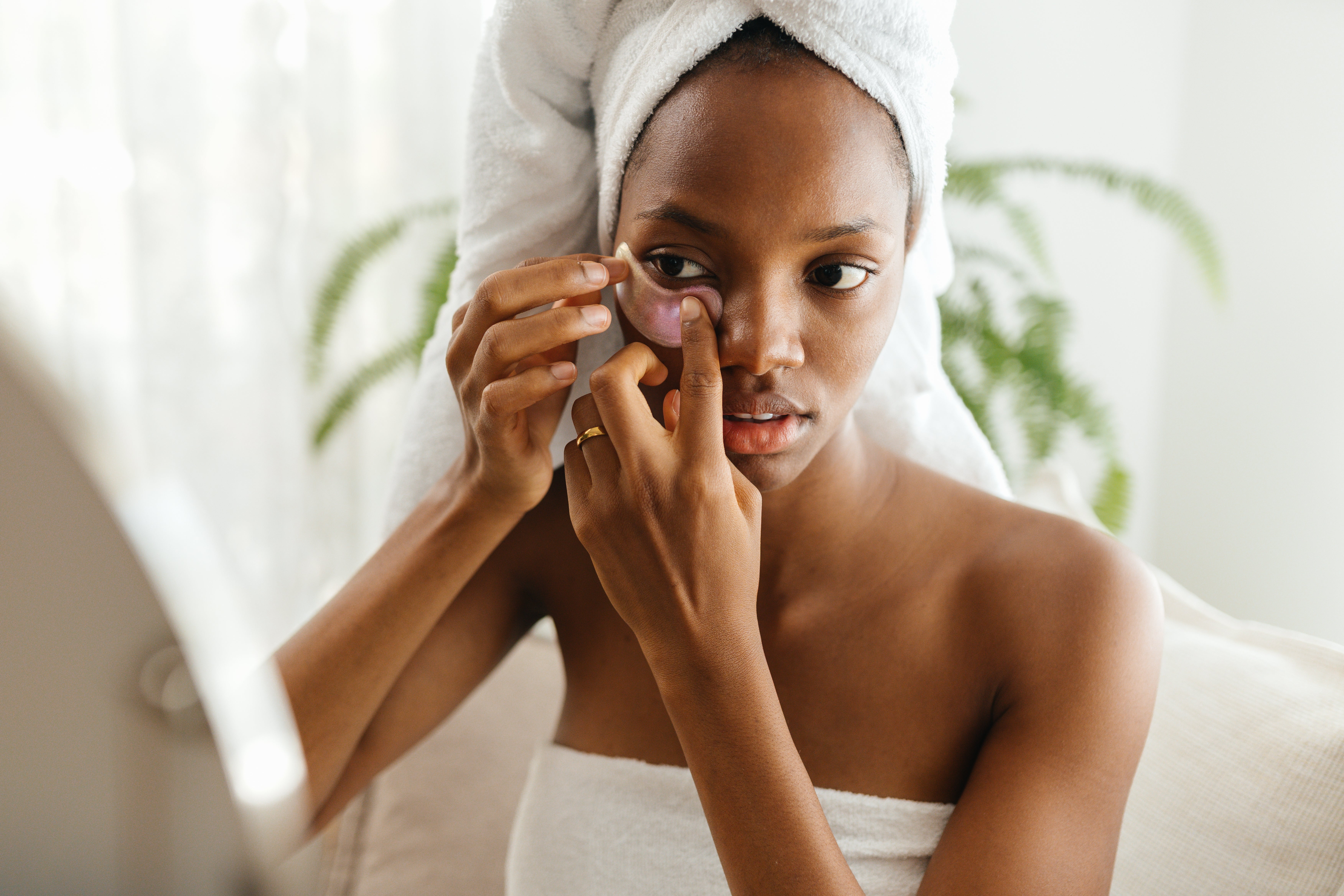 A idiosyncratic with a towel wrapped astir their head, applying a skincare spot nether their oculus portion looking into a mirror