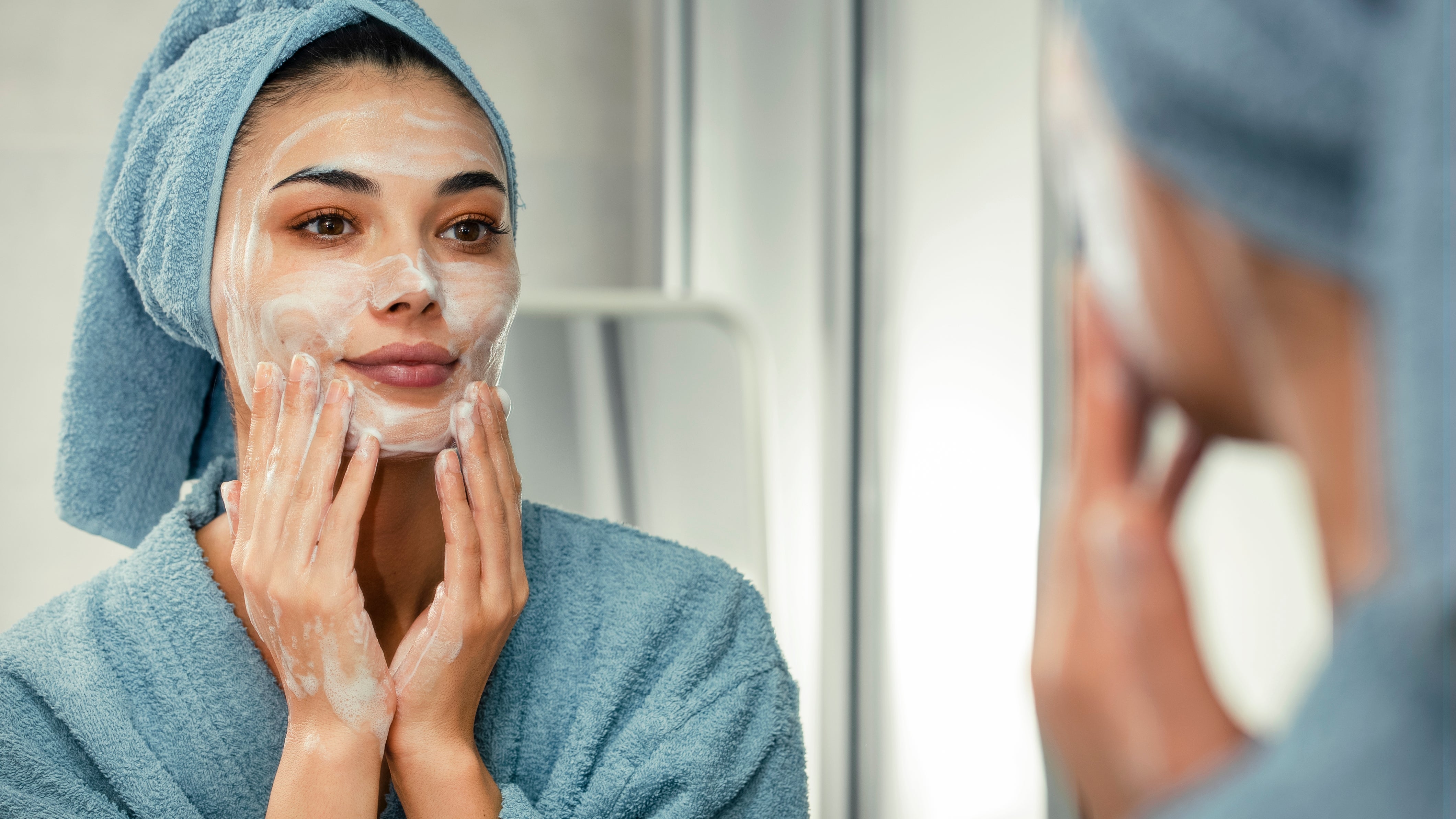 A idiosyncratic wearing a robe and towel astir their head, applying skincare portion looking successful a mirror