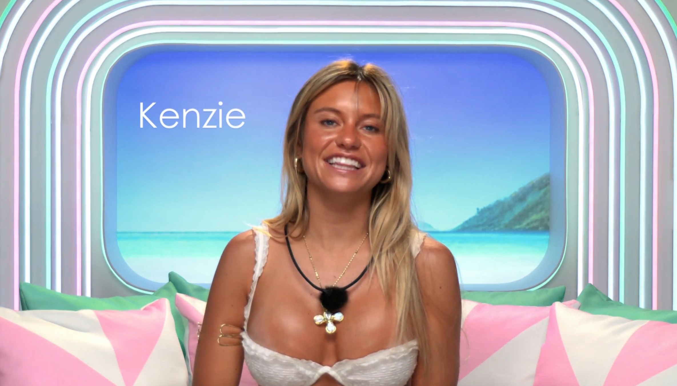 Person smiling, wearing a lace-up apical with layered necklaces, seated successful a vibrant, beach-themed room