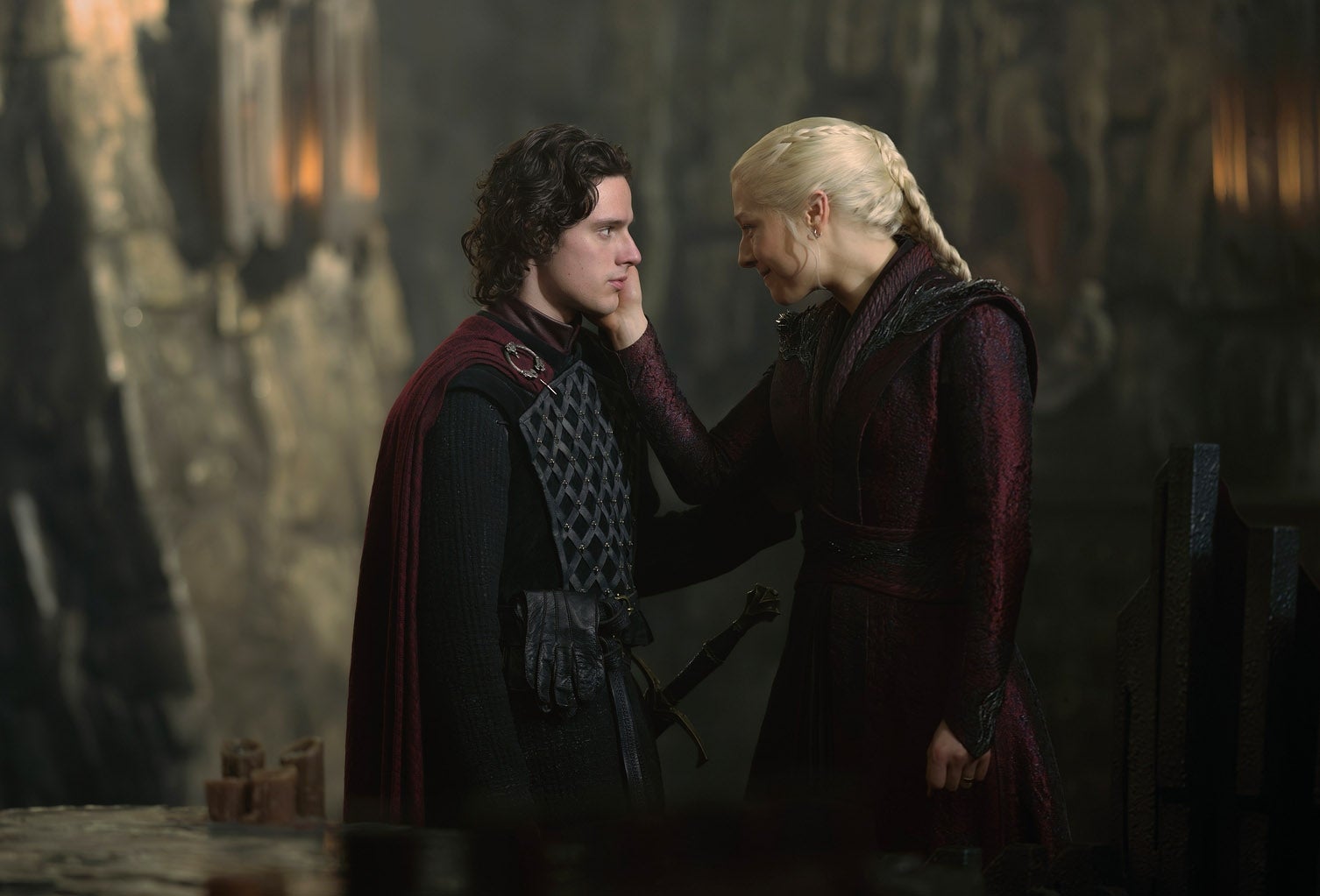 Two characters in medieval-style outfits share an emotional moment in a dimly lit setting, one gently touching the other's face
