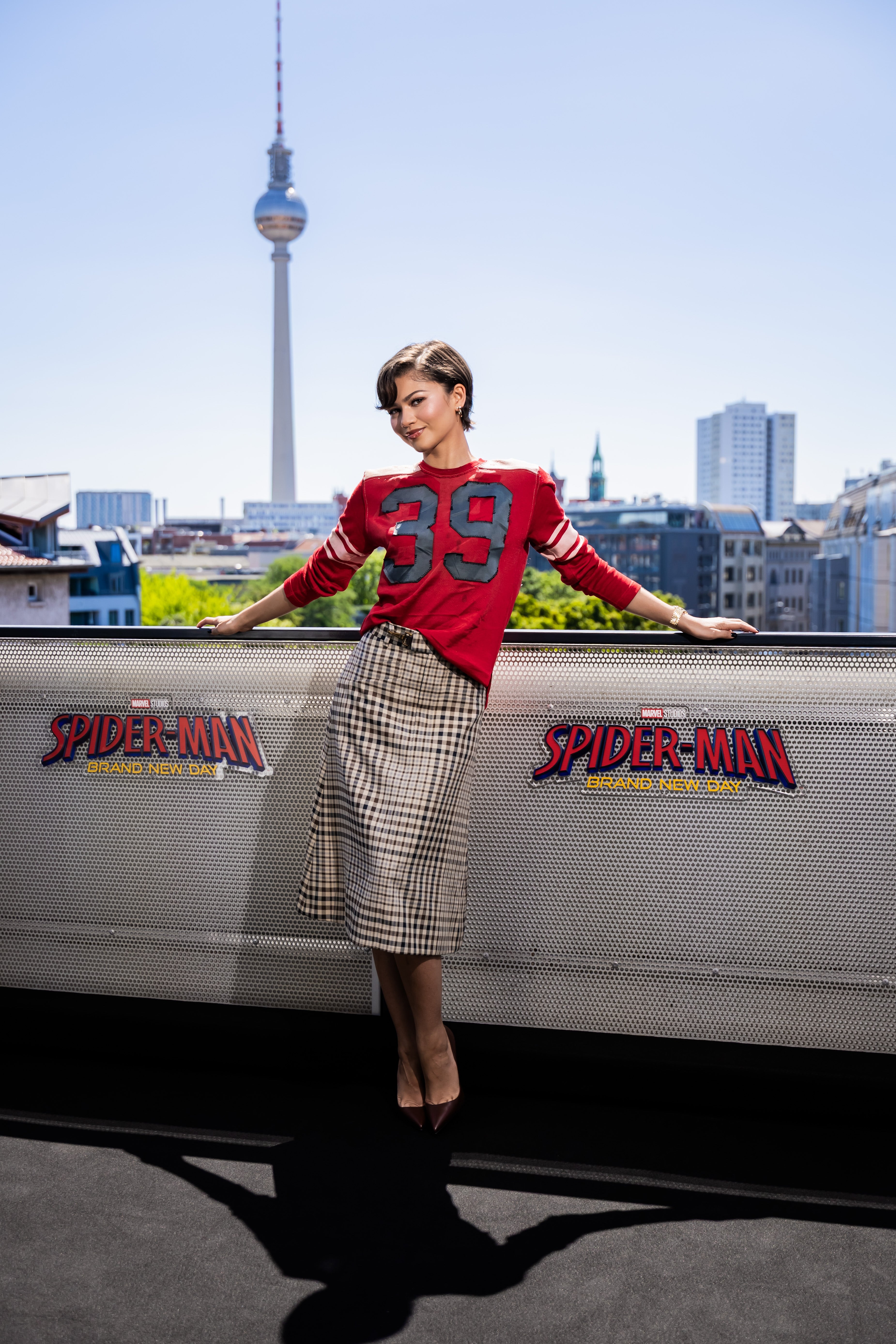 Zendaya posing with a city skyline backdrop, wearing a sweater with the number 39 and a plaid skirt, next to a "Spider-Man" movie banner