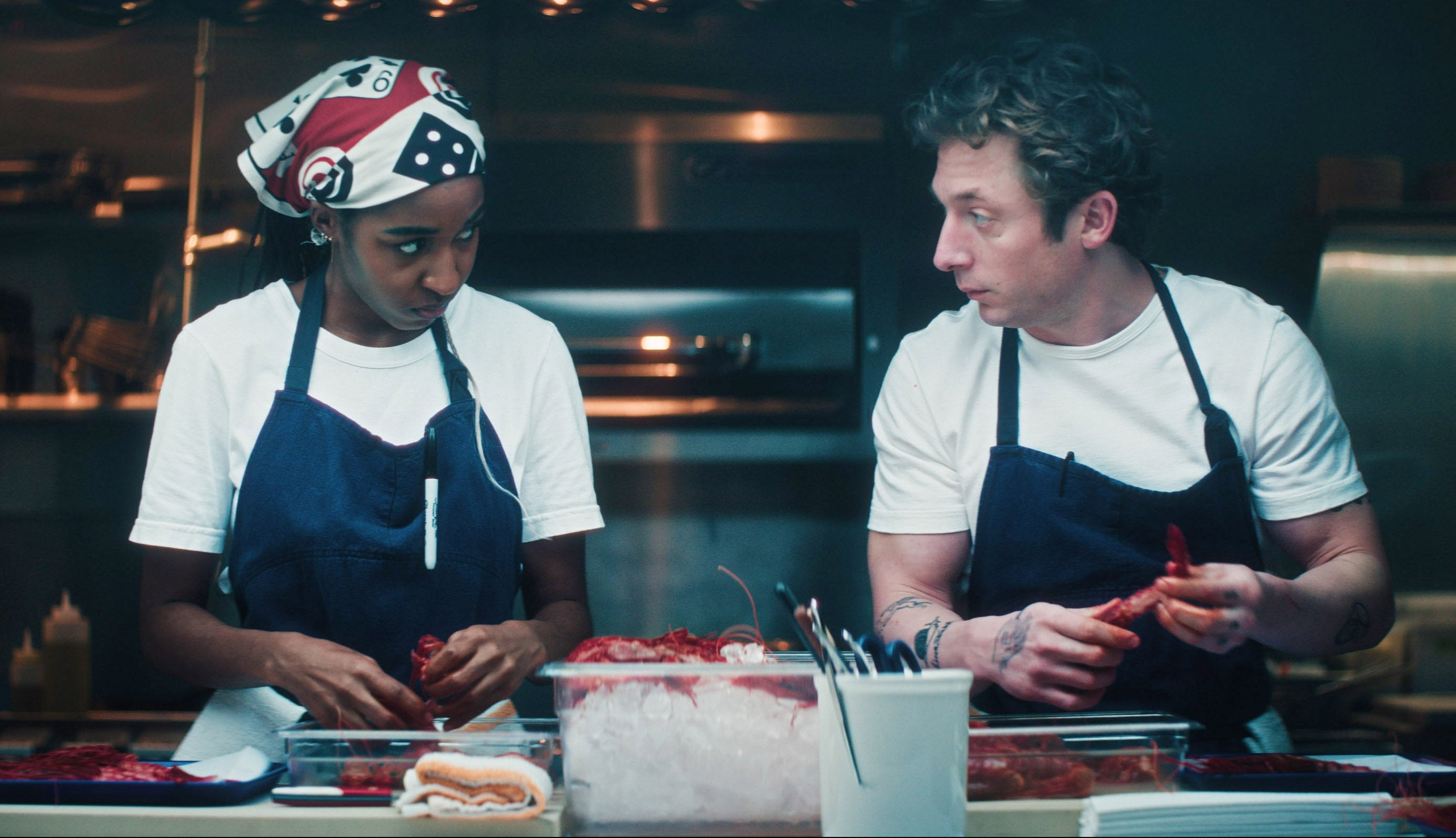 Two chefs in a kitchen, wearing aprons and focused on food preparation. One wears a bandana and the other has tattoos