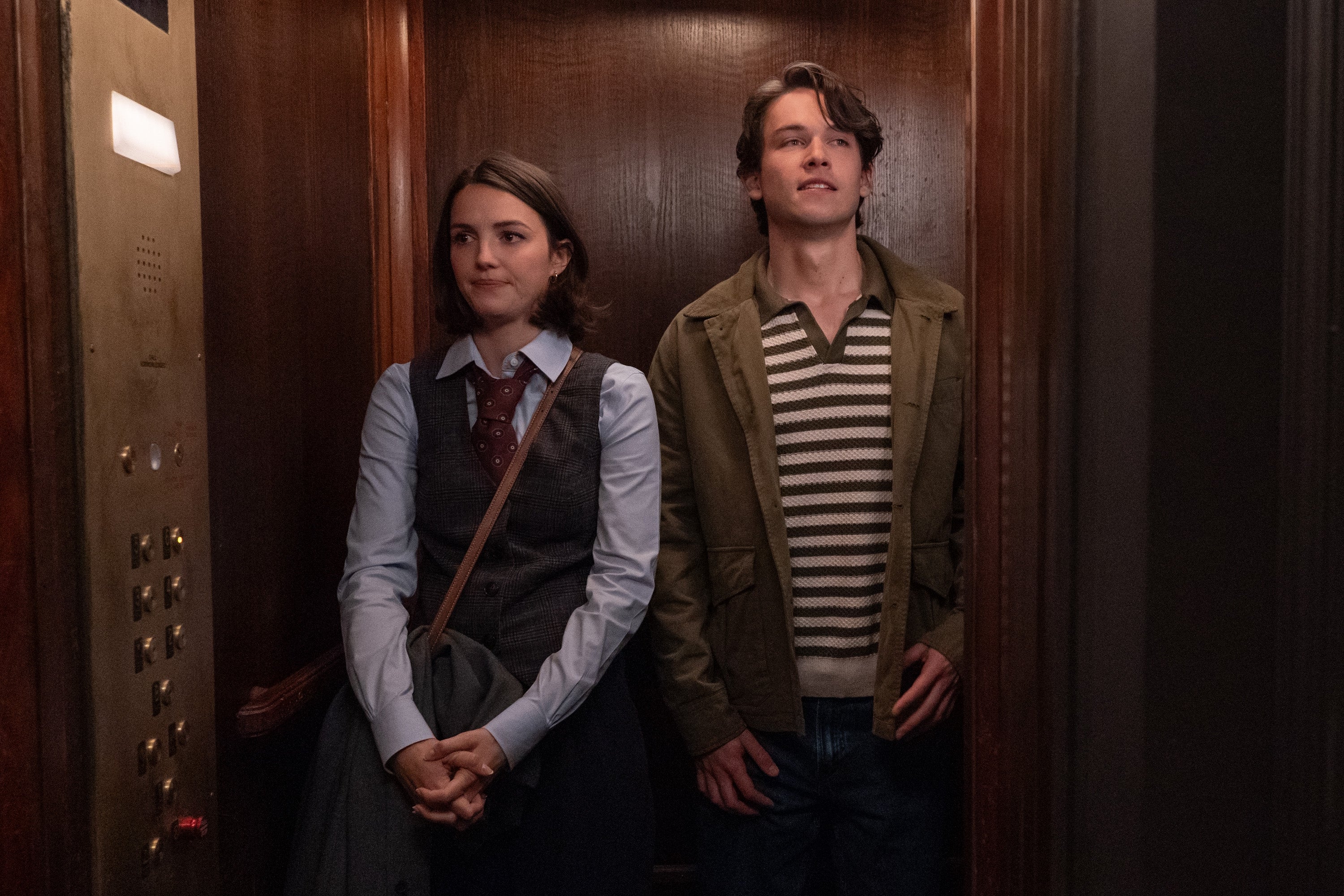 Two individuals stand in an elevator. One wears a vest over a collared shirt and tie; the other wears a striped shirt with a jacket