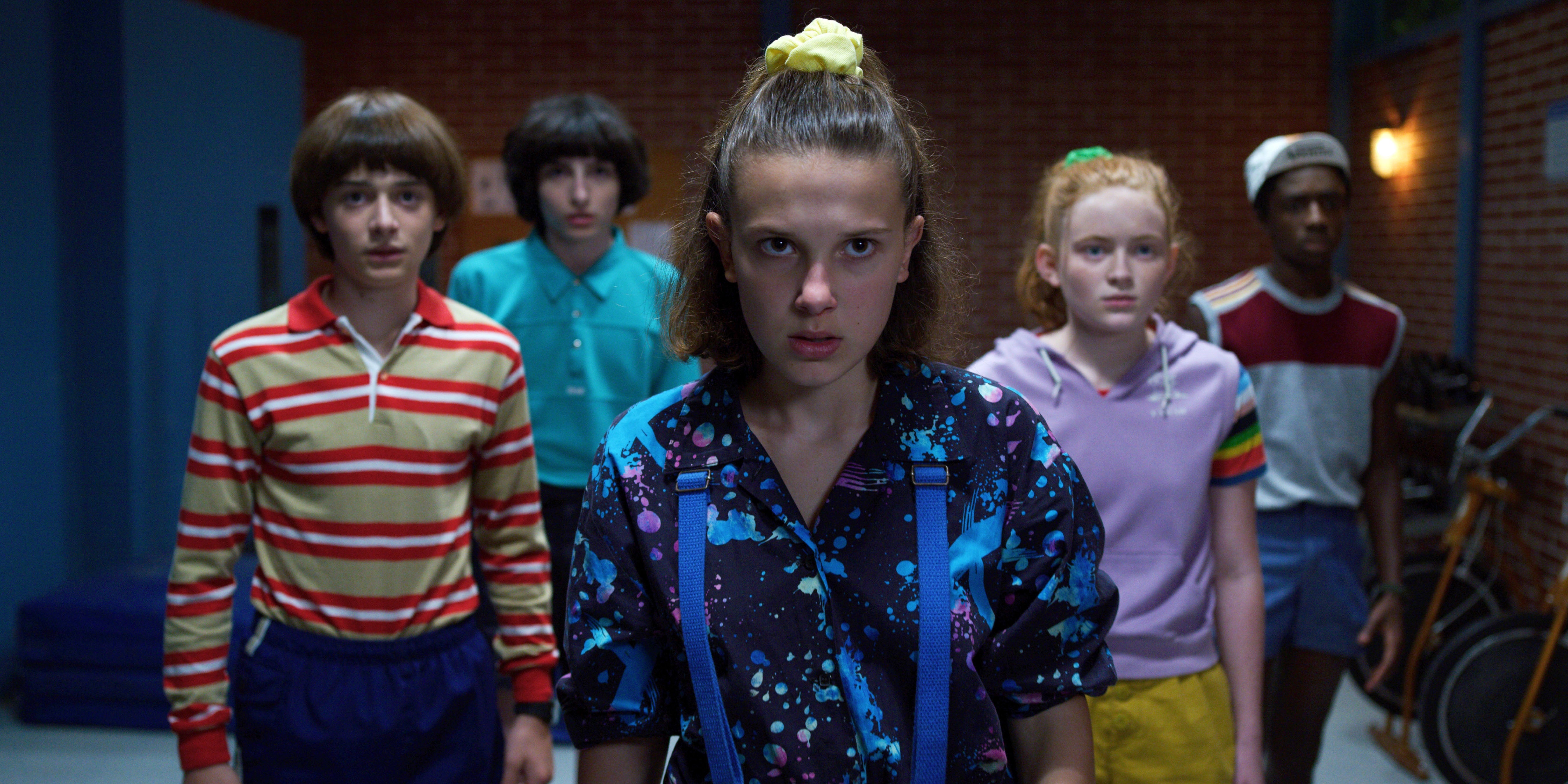 Five teens in retro 80s attire, including bold patterns and stripes, stand together, appearing focused, set in a vintage-inspired indoor background