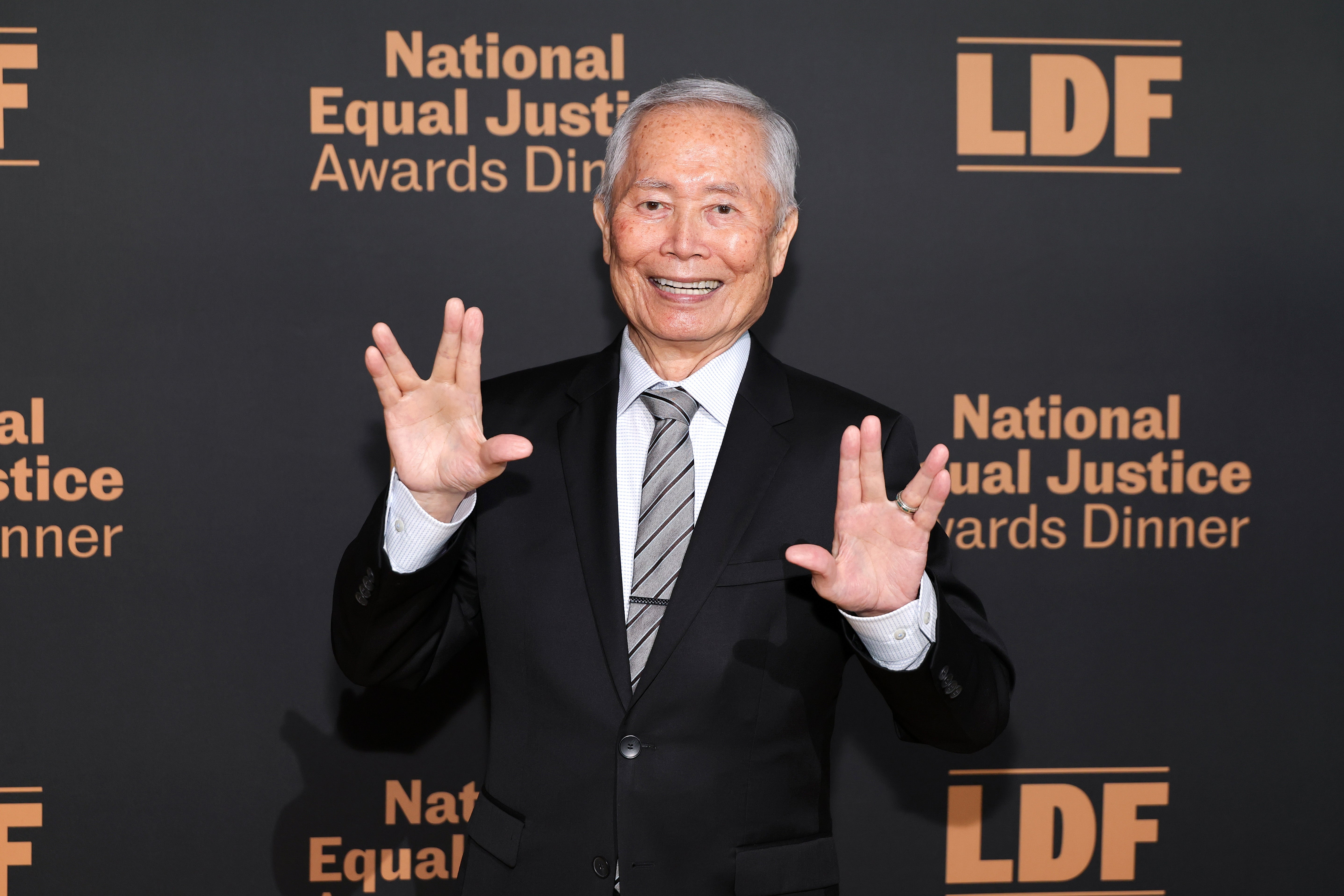 Person posing with a lukewarm grin and motion astatine an awards dinner, wearing a suit and tie, successful beforehand of an lawsuit backdrop