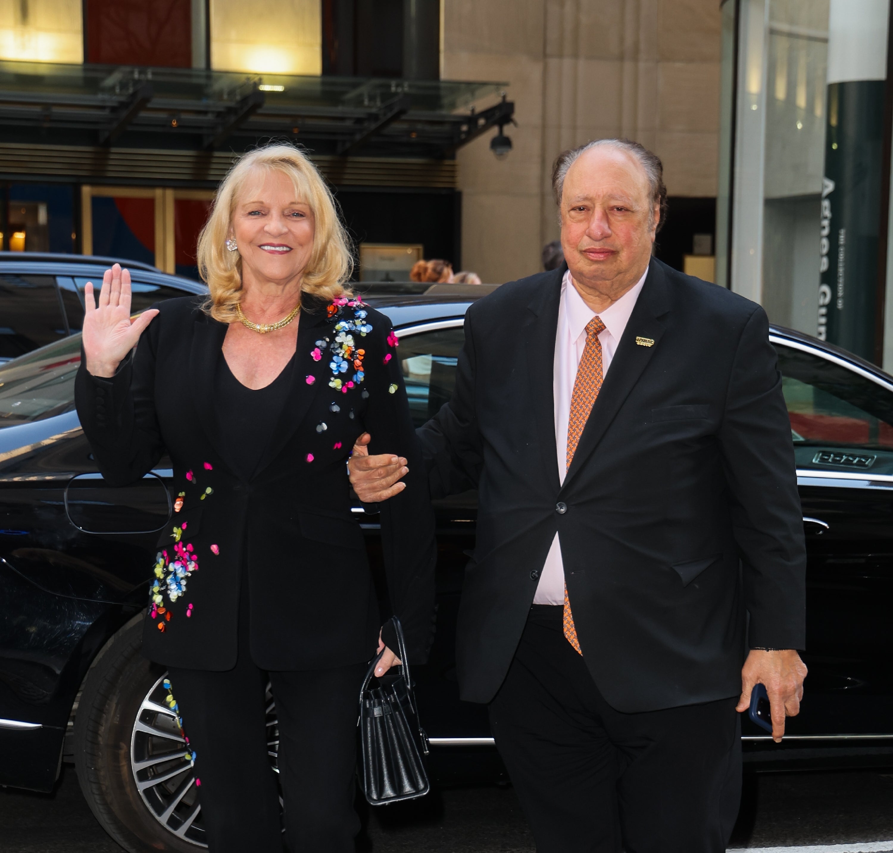 Two radical smiling and waving outside, 1 successful a floral blazer and the different successful a suit, lasting beside a car
