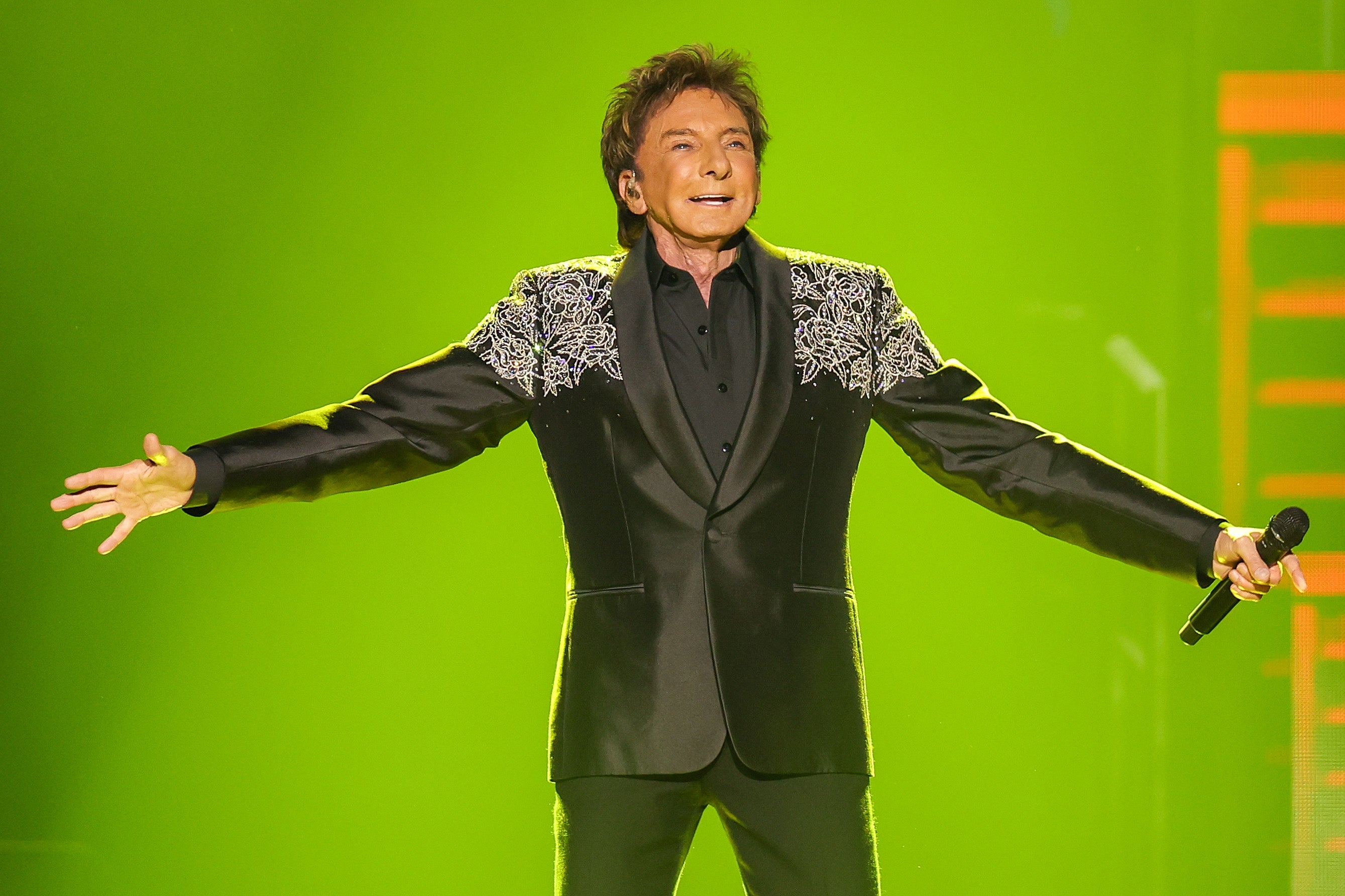 Person singing connected stage, wearing a achromatic suit with intricate enarthrosis details, holding a microphone. Arms outstretched, expressing vigor and joy