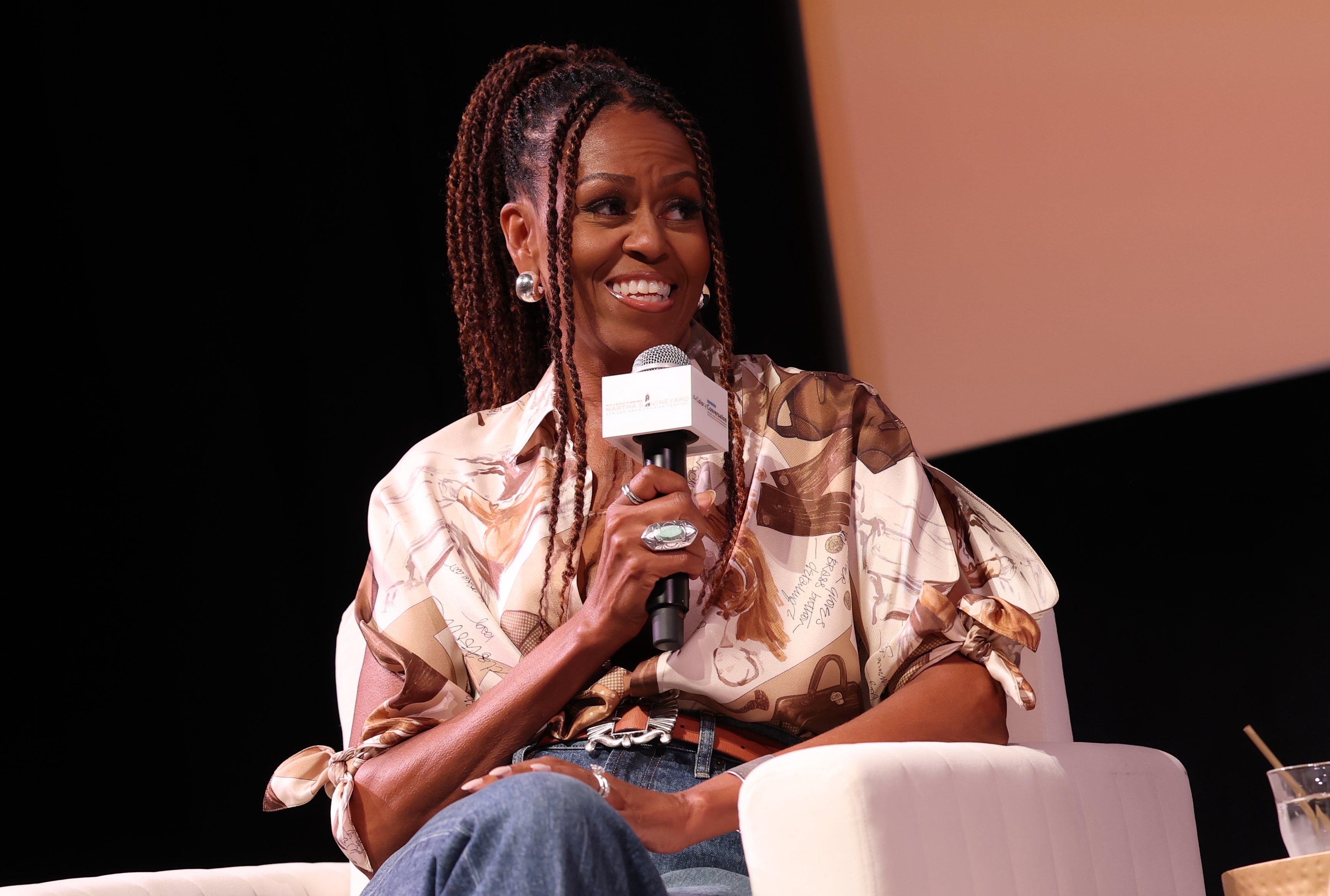 A idiosyncratic with braided hairsbreadth wearing a patterned garment and jeans sits connected signifier during a discussion, smiling and holding a microphone