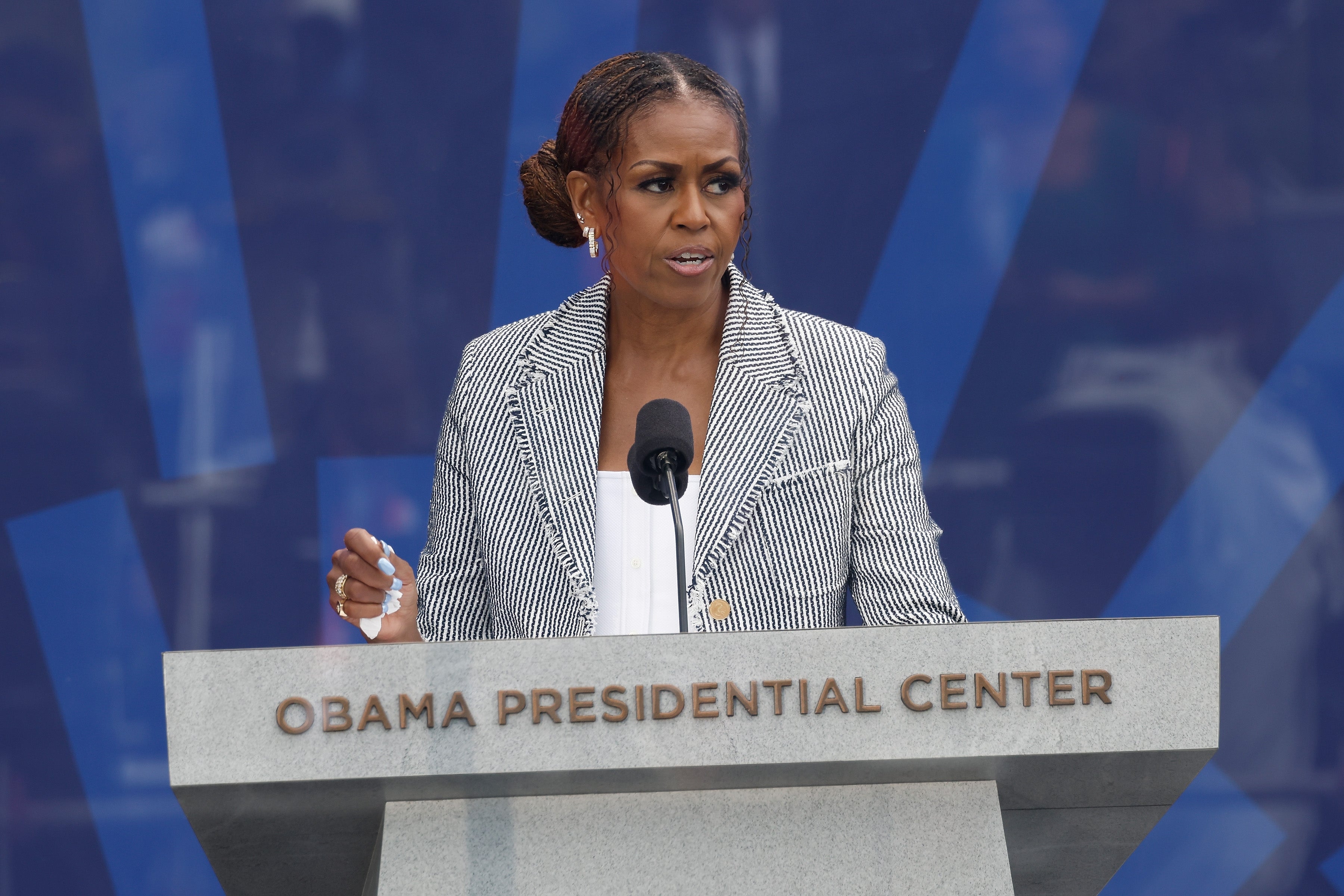 Person speaking astatine a podium labeled "Obama Presidential Center," wearing a striped blazer and achromatic top