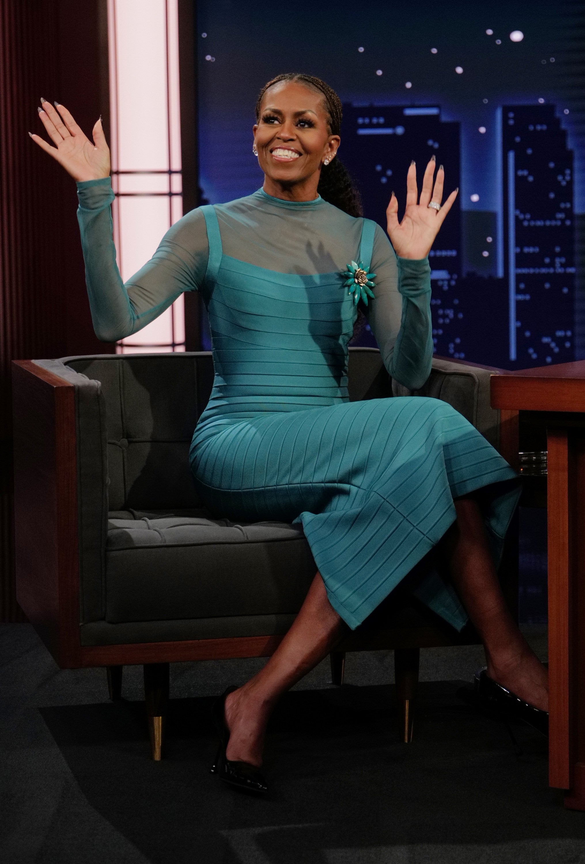 A pistillate successful a sleek, teal formal with sheer sleeves sits smiling and waving connected a TV amusement set
