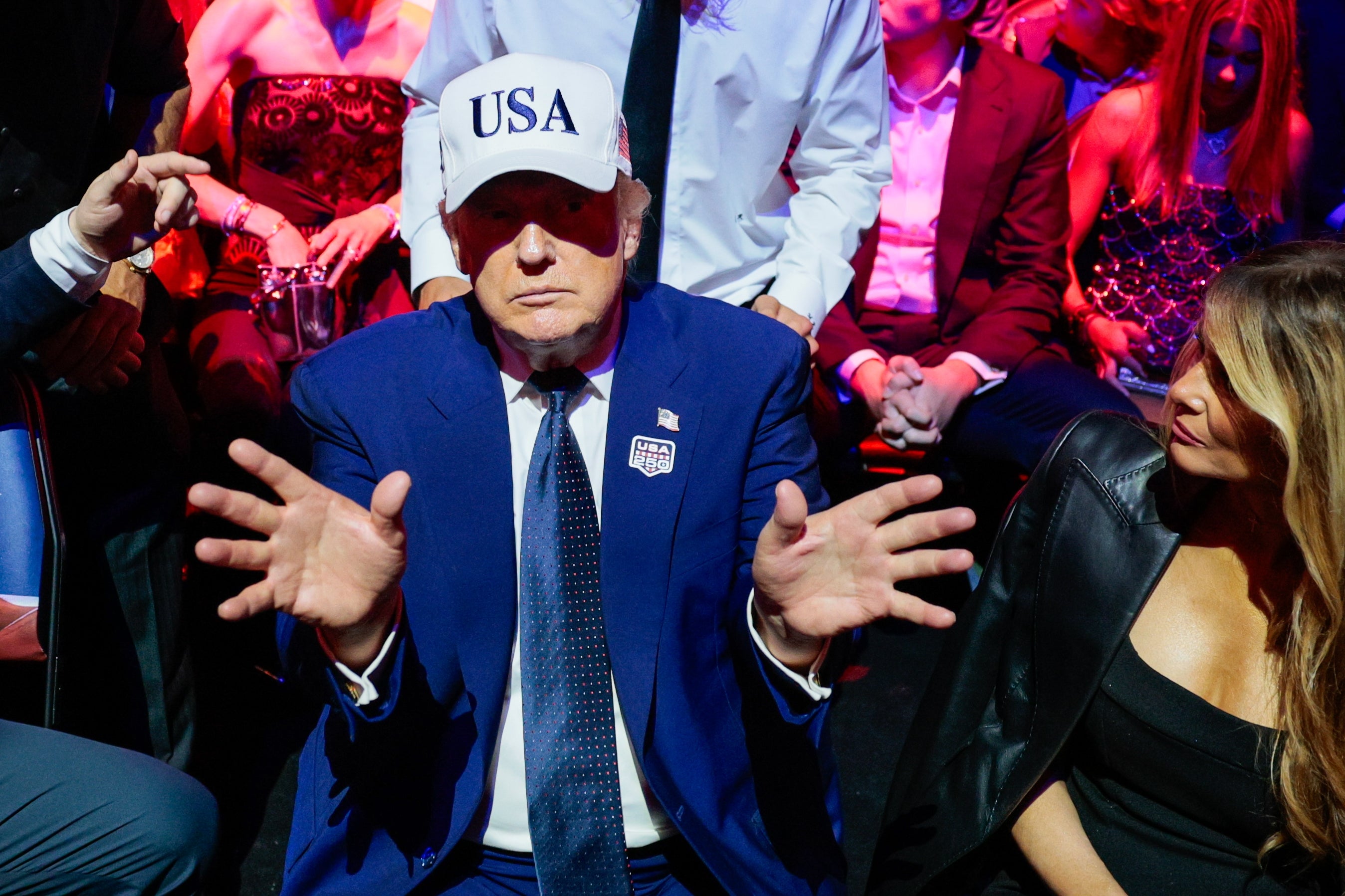 A idiosyncratic successful a suit and a "USA" headdress is seated, gesturing with unfastened hands, surrounded by others astatine an indoor event