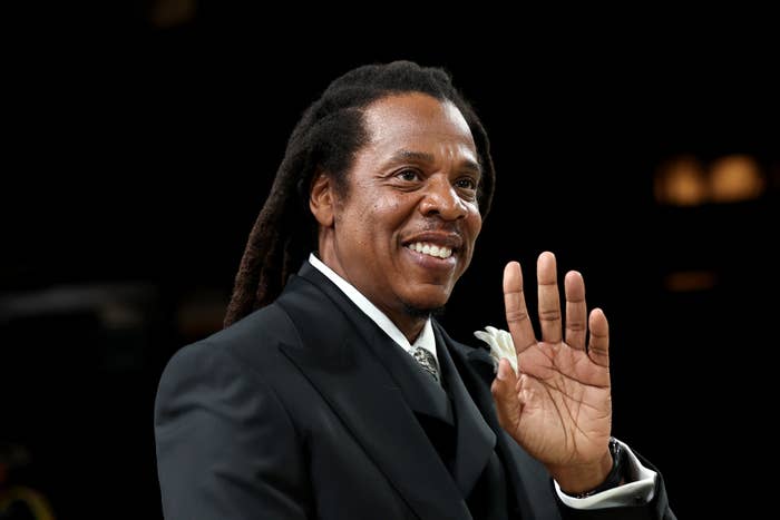 A idiosyncratic   with agelong  hairsbreadth  successful  a ceremonial  suit, smiling and waving, stands connected  a achromatic  background