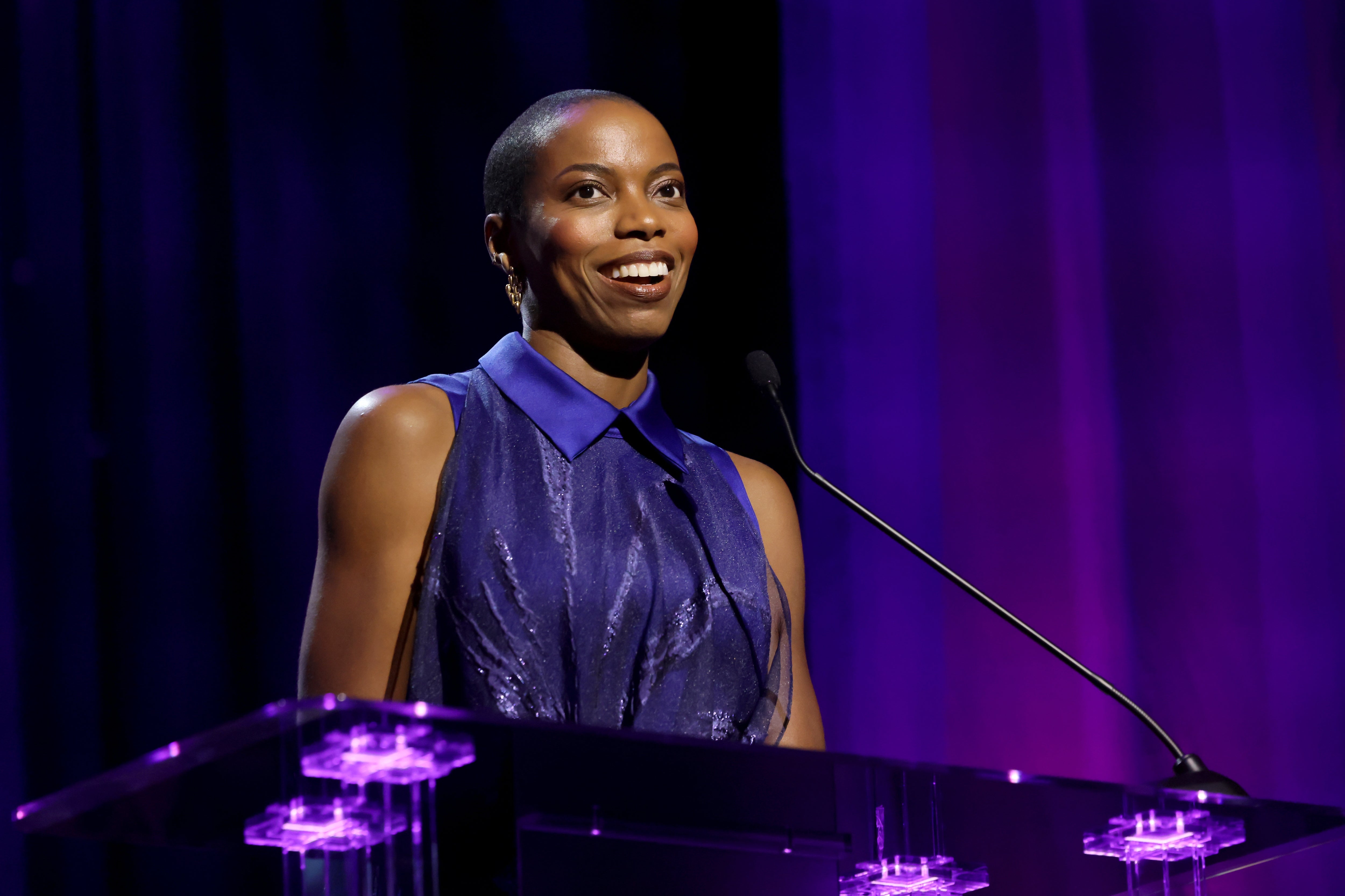 Person smiling and speaking astatine a podium, wearing a sleeveless, shimmering outfit