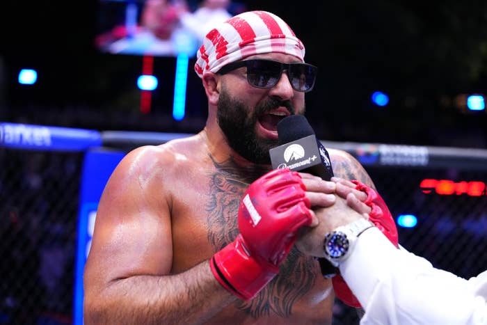 A muscular antheral   with a beard and tattoos speaks into a microphone, wearing a striped bandana, sunglasses, and gloves, successful  a ringing  oregon  octagon setting