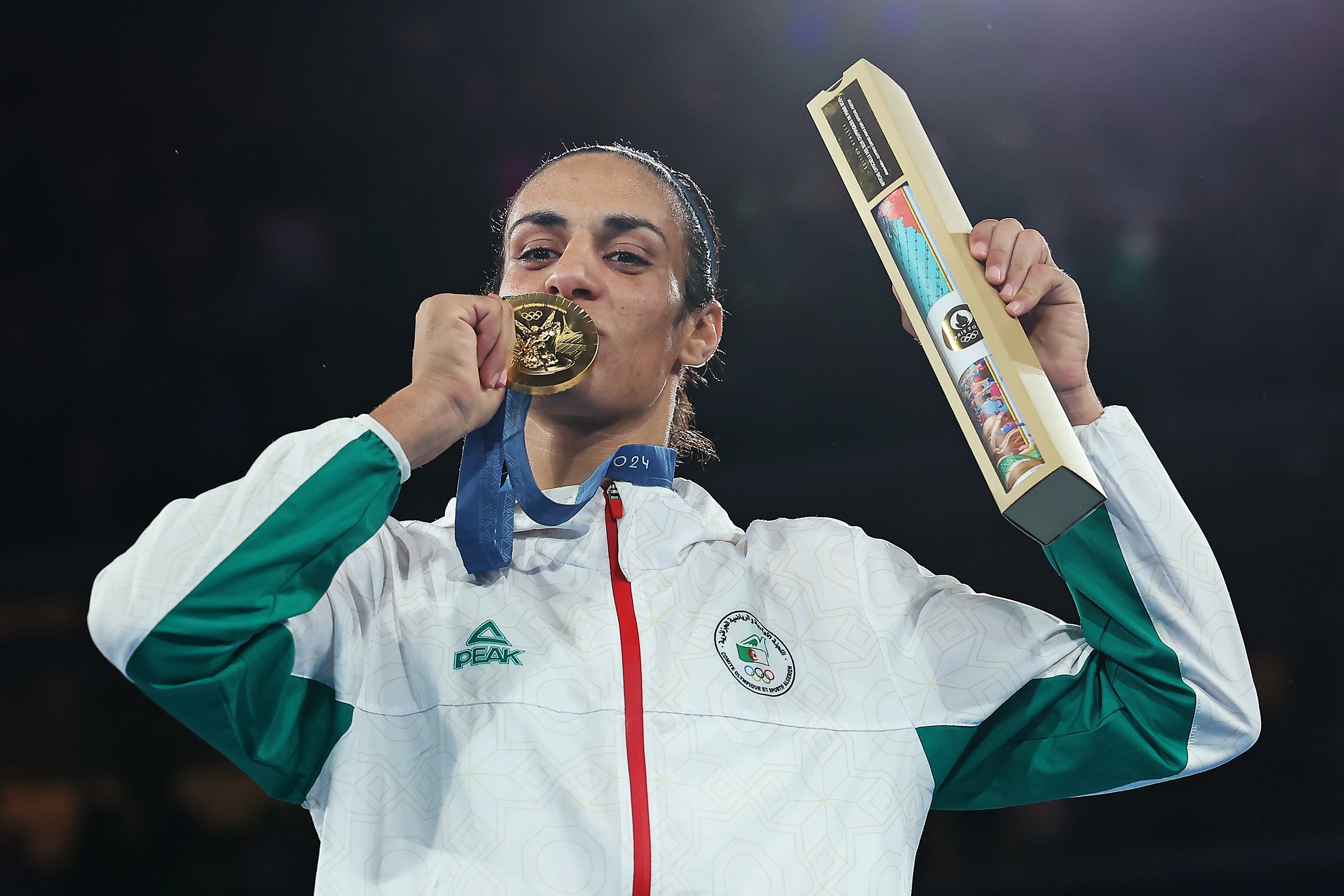 Athlete kisses golden medal, holding a trophy, wearing a sports overgarment with an Olympic logo