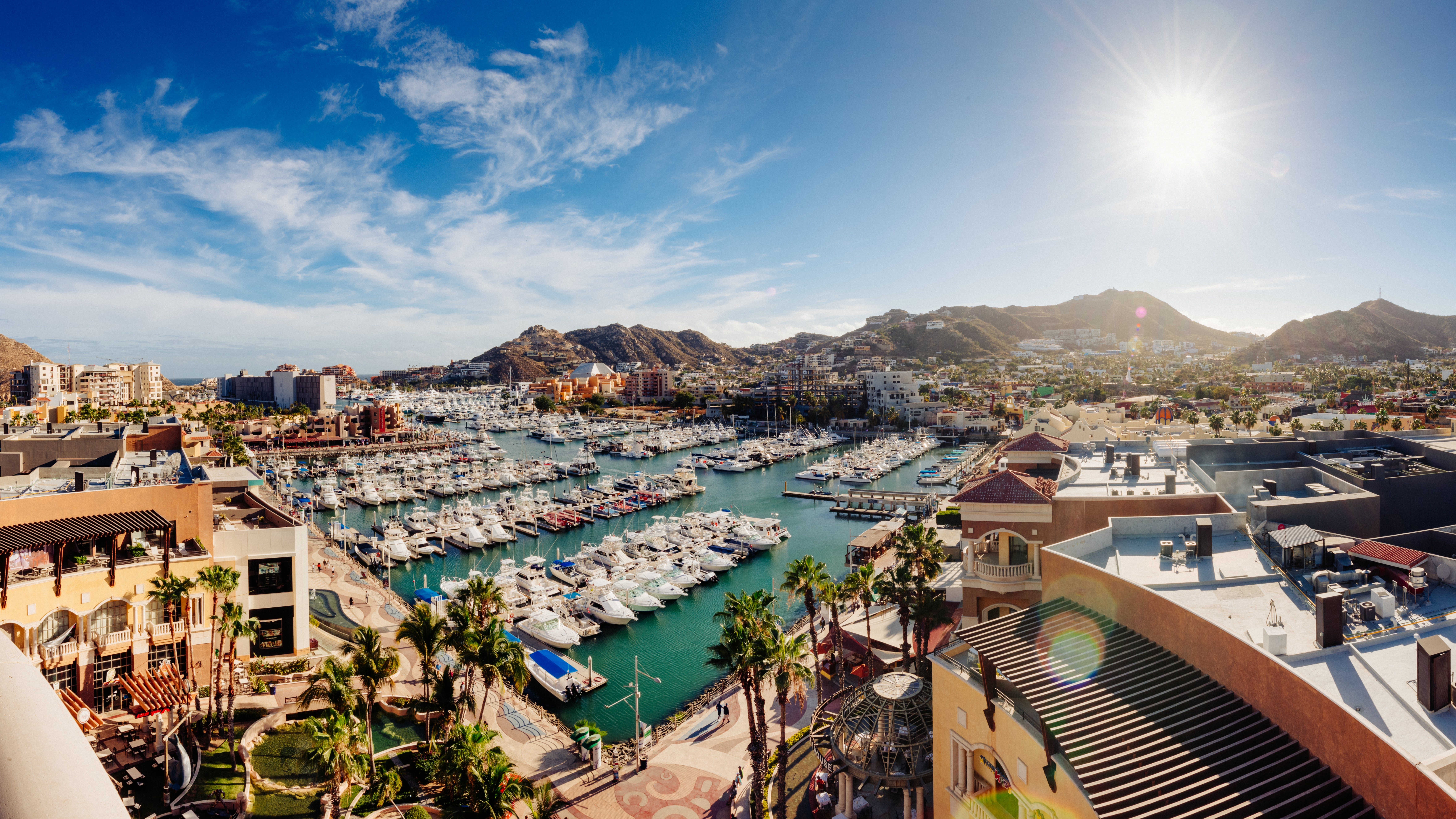 View of a marina with galore boats docked, surrounded by buildings and mountains nether a sunny sky