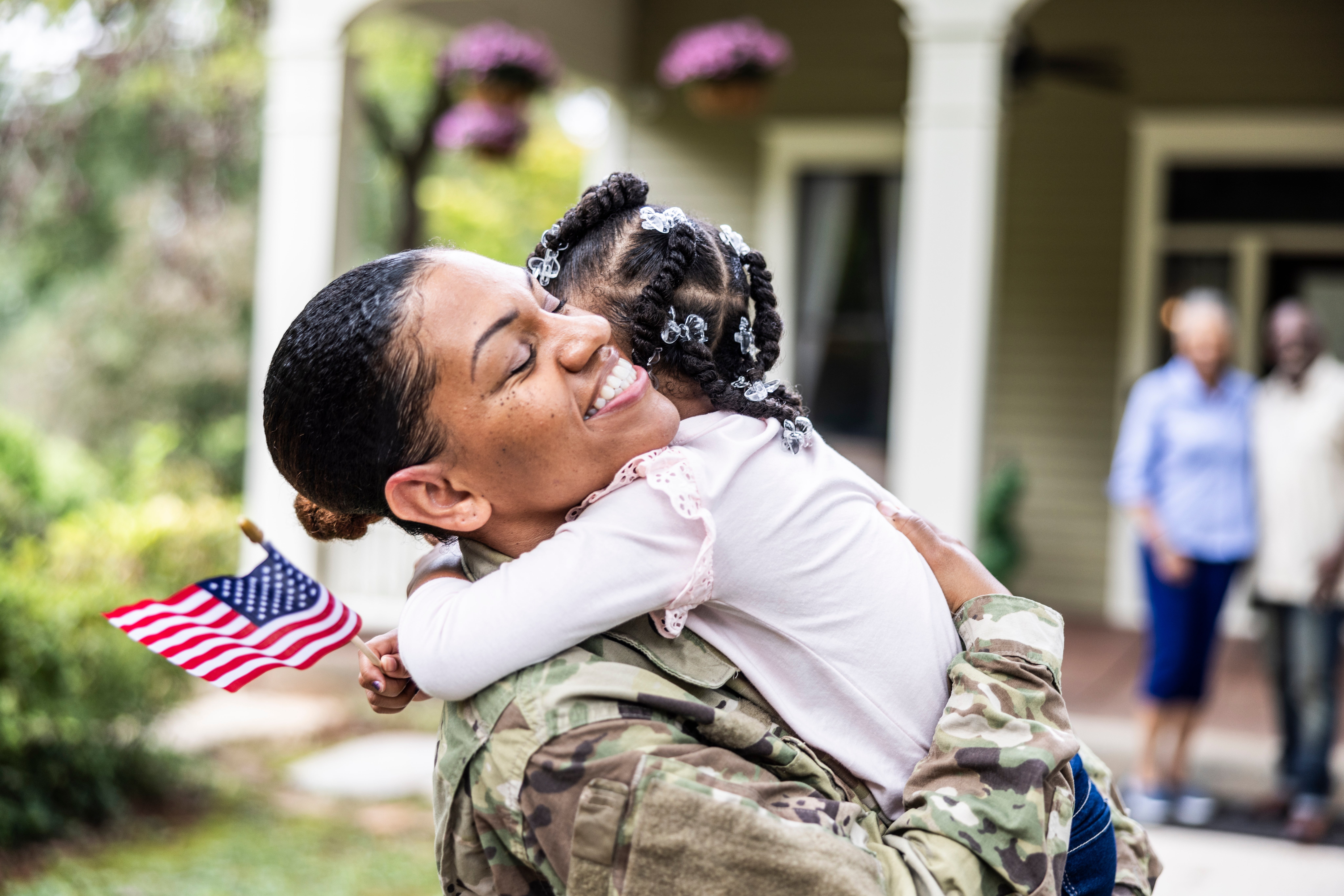 A smiling worker  successful  camouflage embraces a kid  holding an American flag, with household  successful  the inheritance  extracurricular  a home