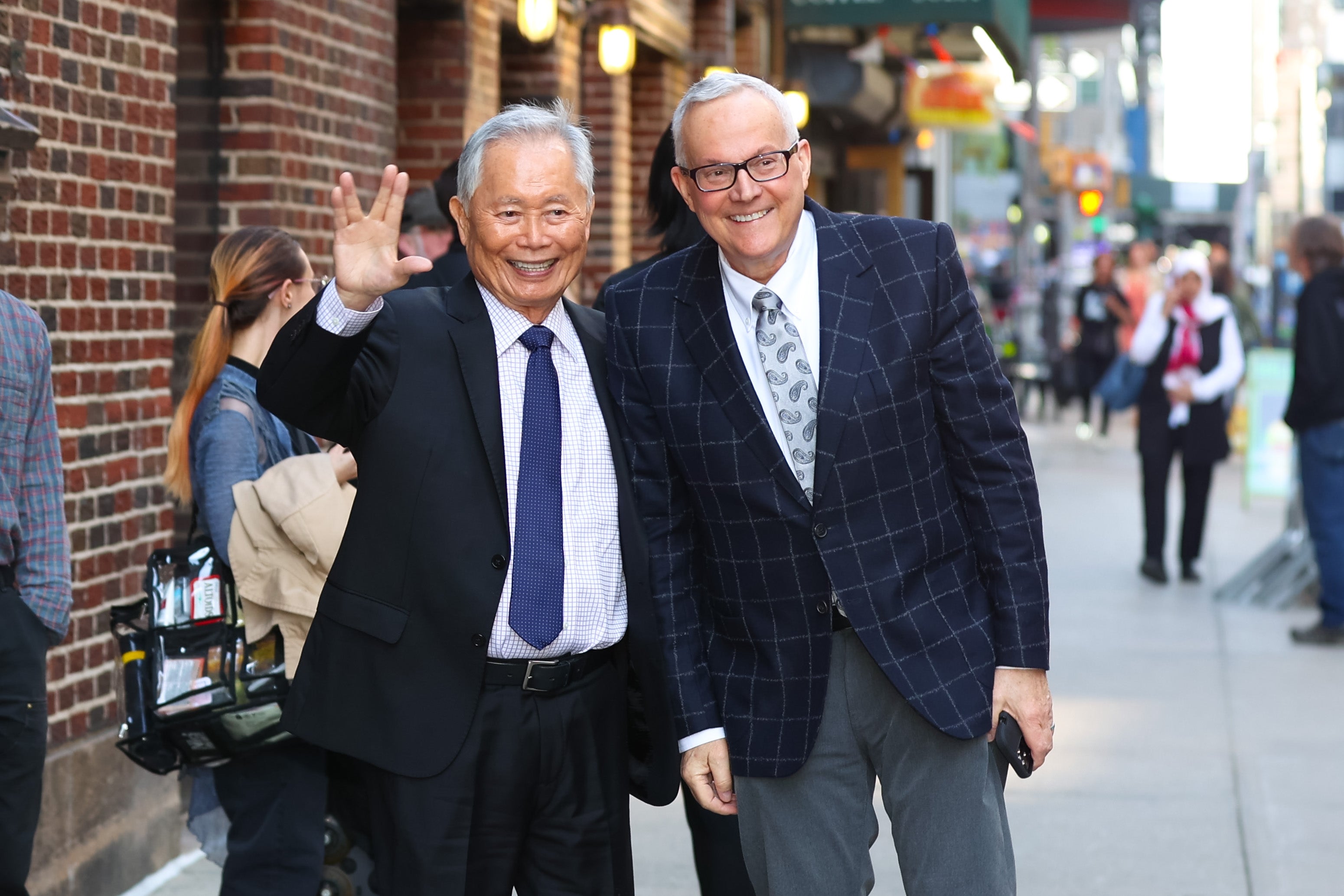 Two radical are smiling and posing connected a metropolis sidewalk. One idiosyncratic is waving with a unsocial manus gesture, and some are wearing suits
