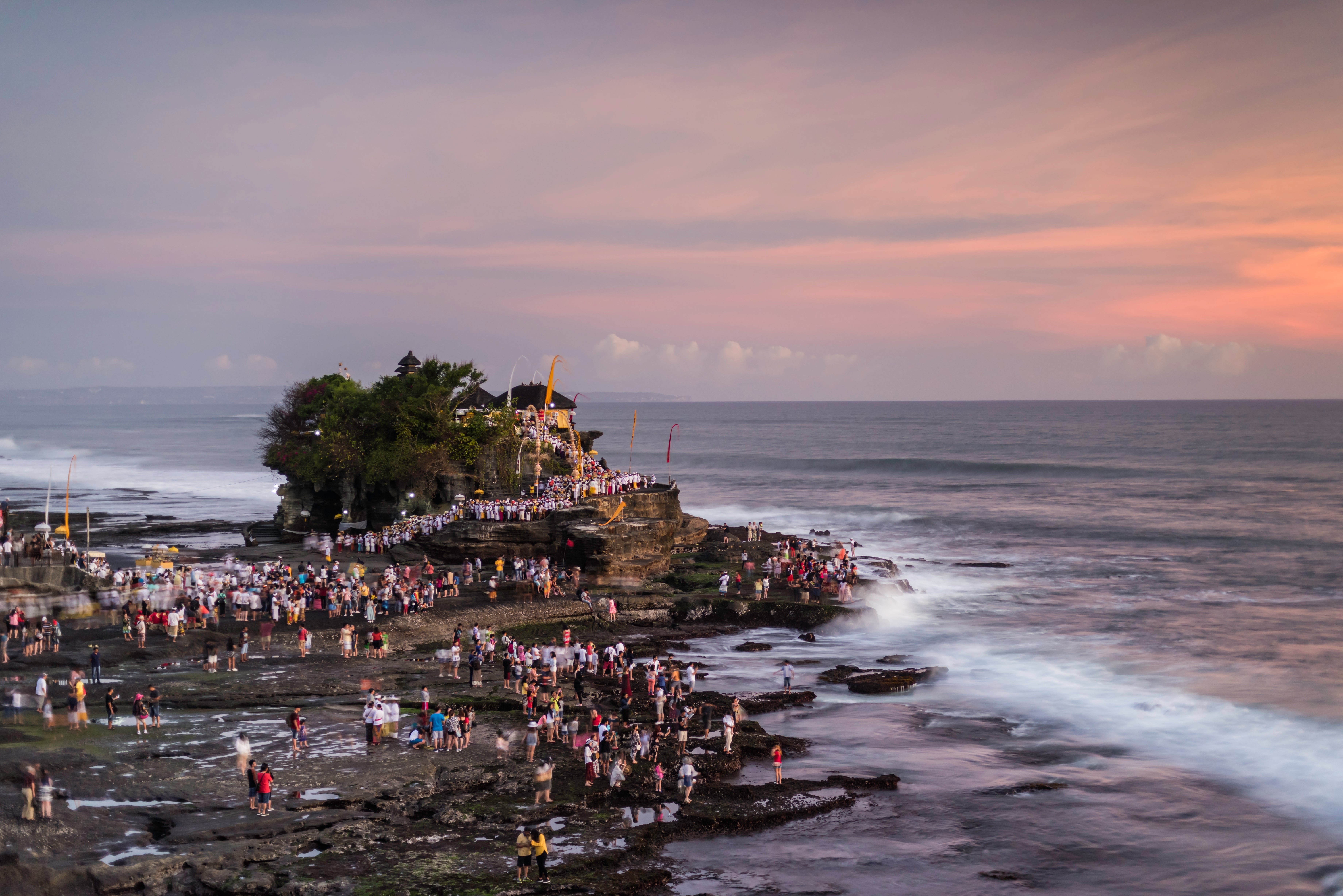 A crowded coastline with radical gathering astatine Tanah Lot Temple successful Bali during sunset, with waves crashing against the rocks