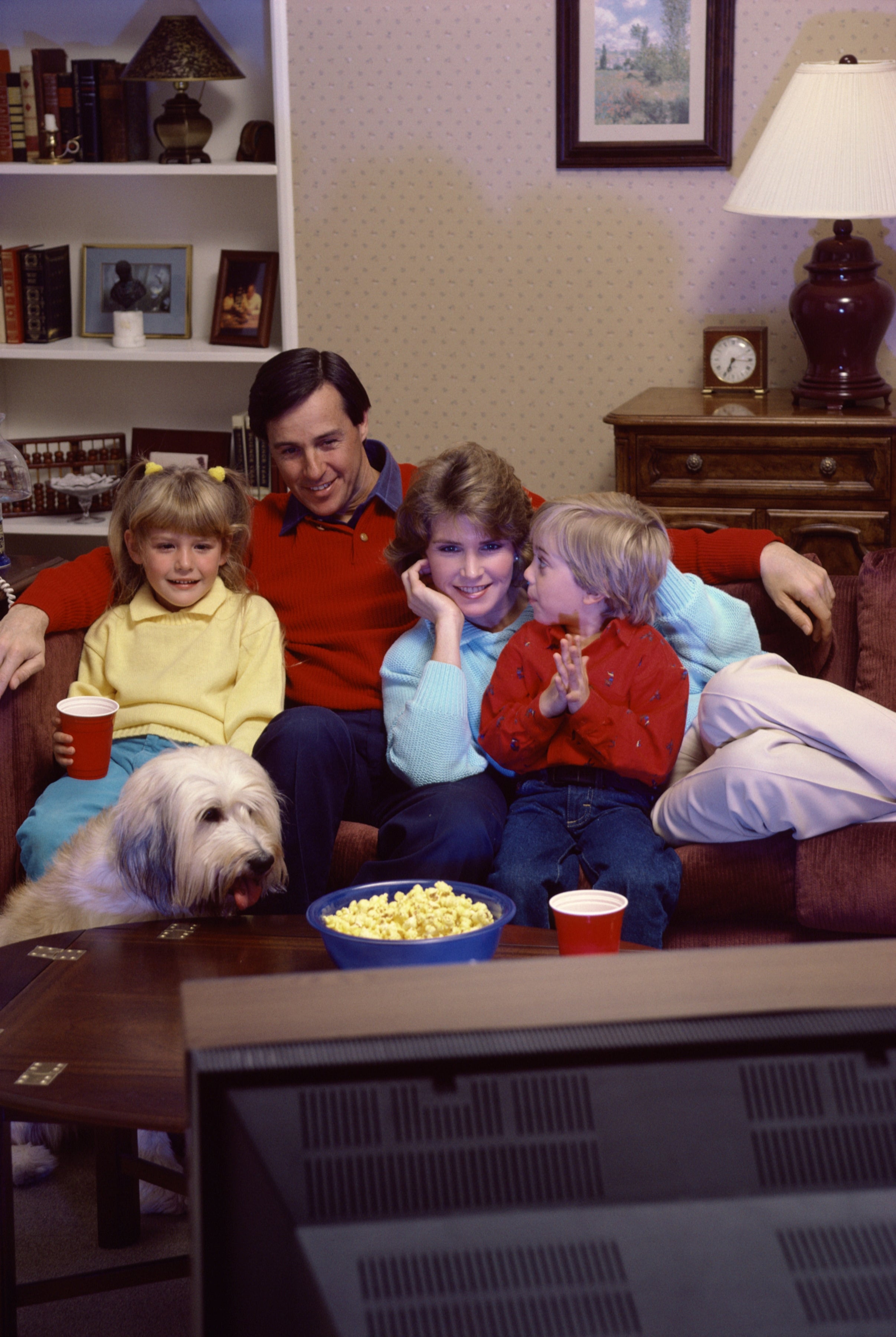 A household  sits connected  a sofa  with a dog, watching TV, surrounded by snacks and drinks, enjoying a cozy evening astatine  home
