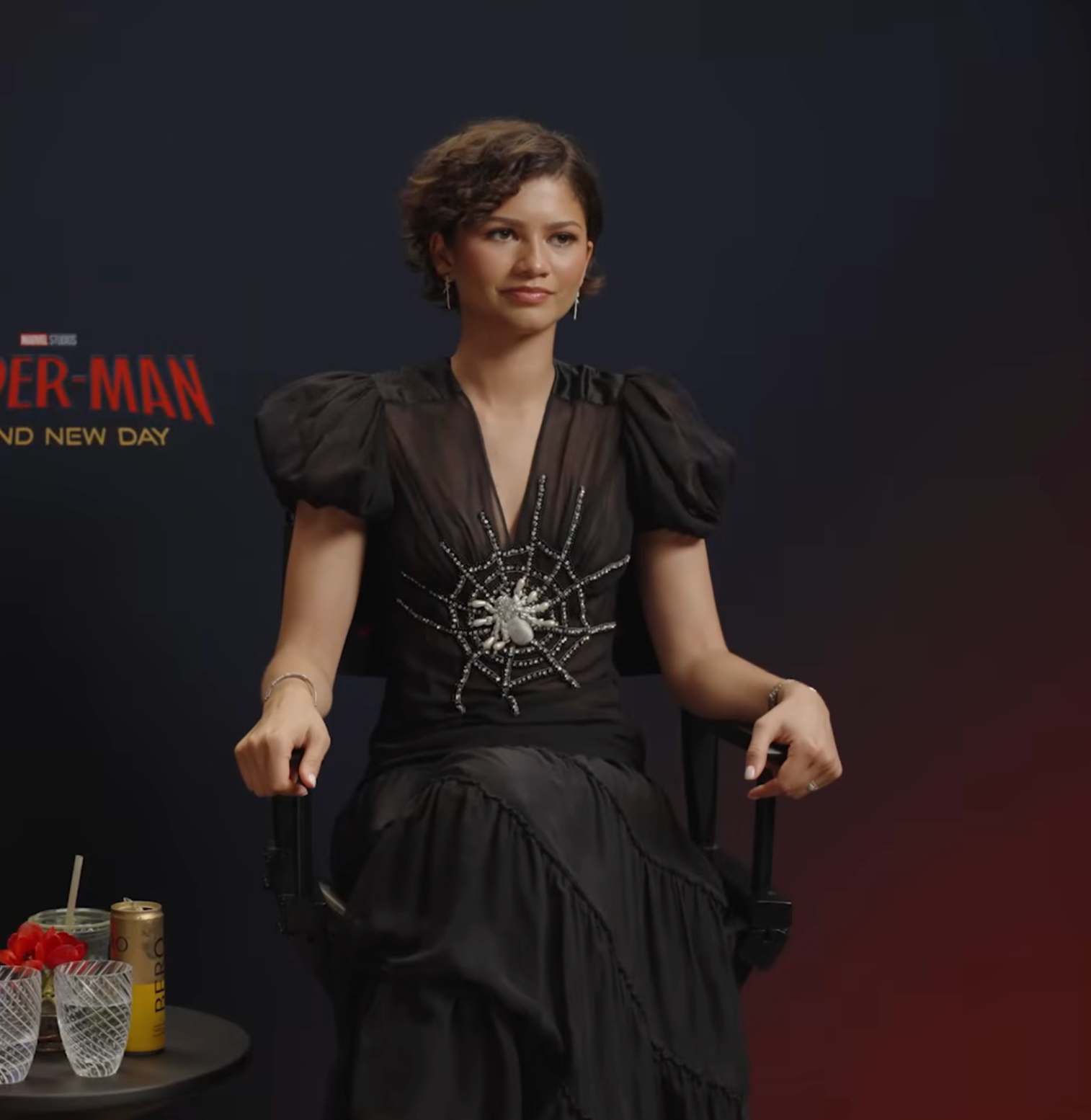Zendaya sits in an interview setting, wearing a stylish black dress with a beaded spider design. They are in front of a movie-themed backdrop
