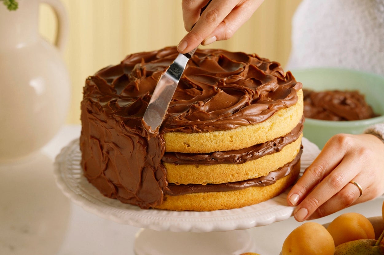 Person frosting a layered cake with chocolate icing on a plate