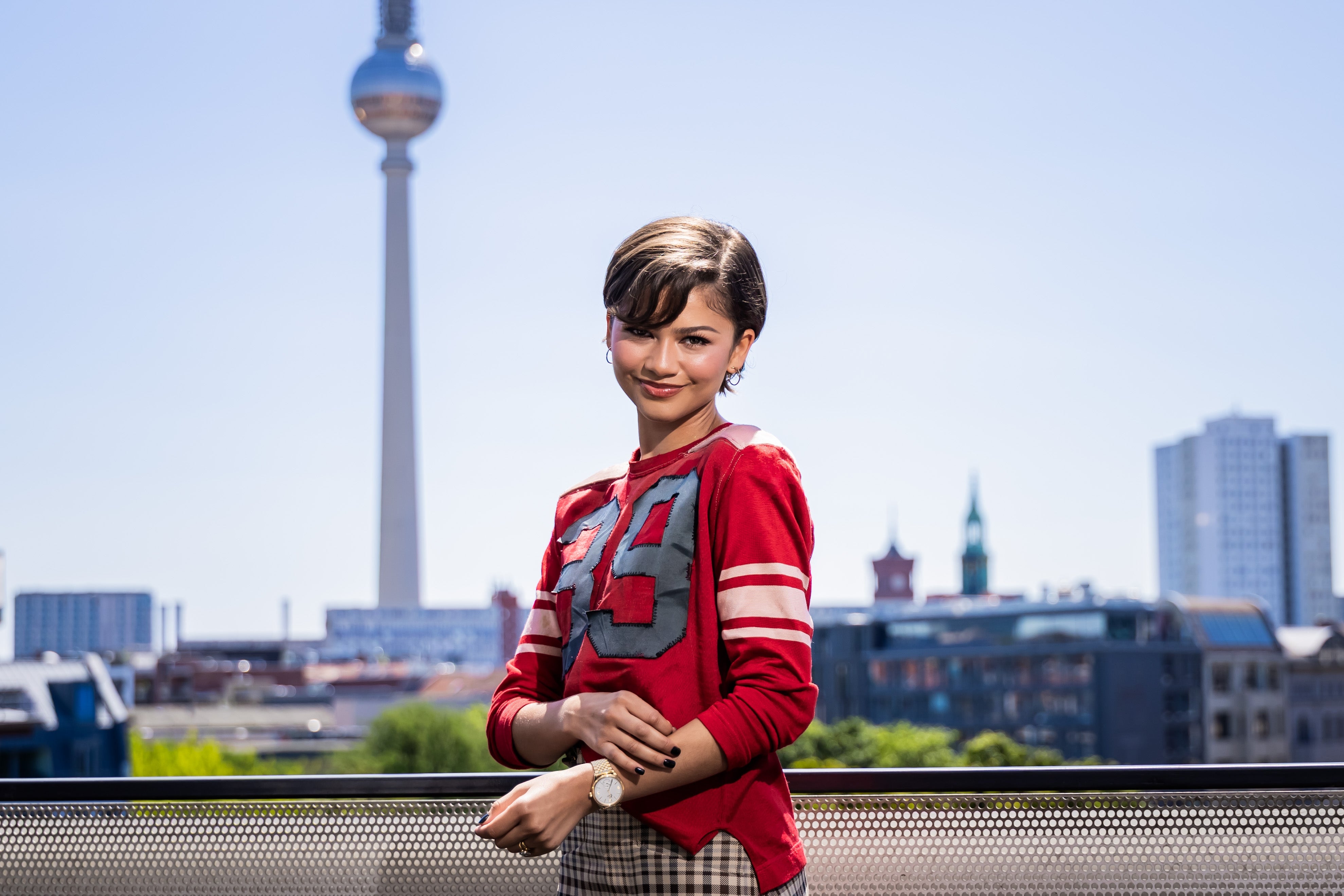 Zendaya in casual sporty outfit with a large number print on the shirt stands outdoors, with a city skyline and a tall tower in the background