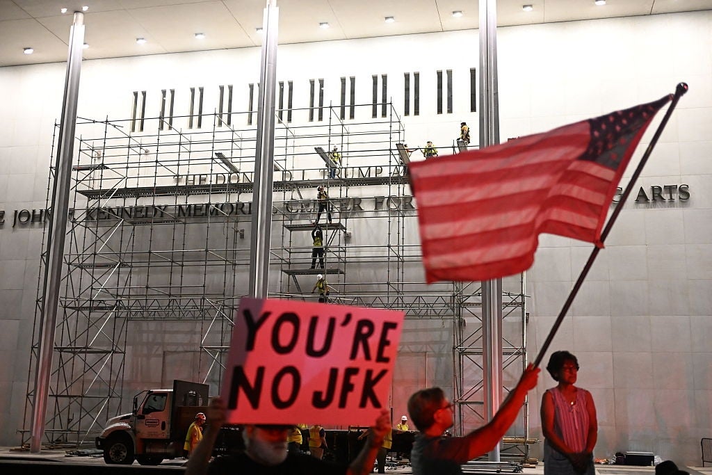 Demonstranter utenfor en bygning med stillas holder et amerikansk flagg og et skilt med teksten