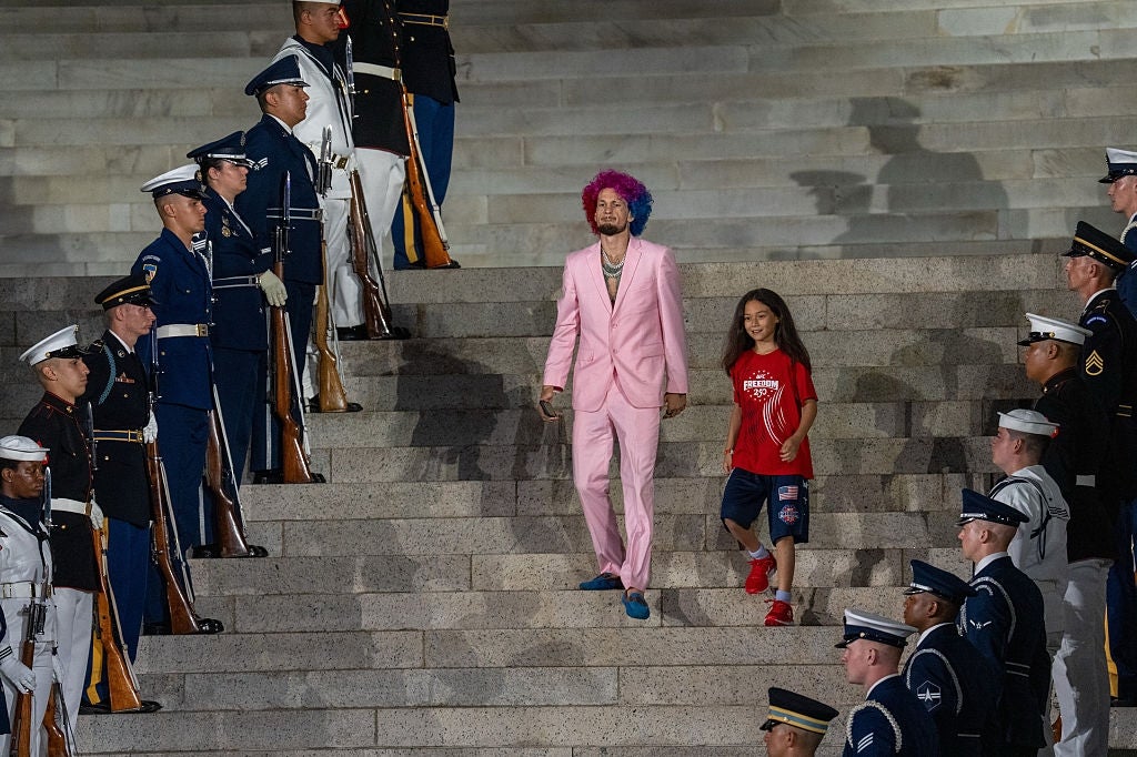 Mann i rosa dress og fargerik parykk, med et barn i uformelle klær, går ned trappene flankert av vakter i uniform