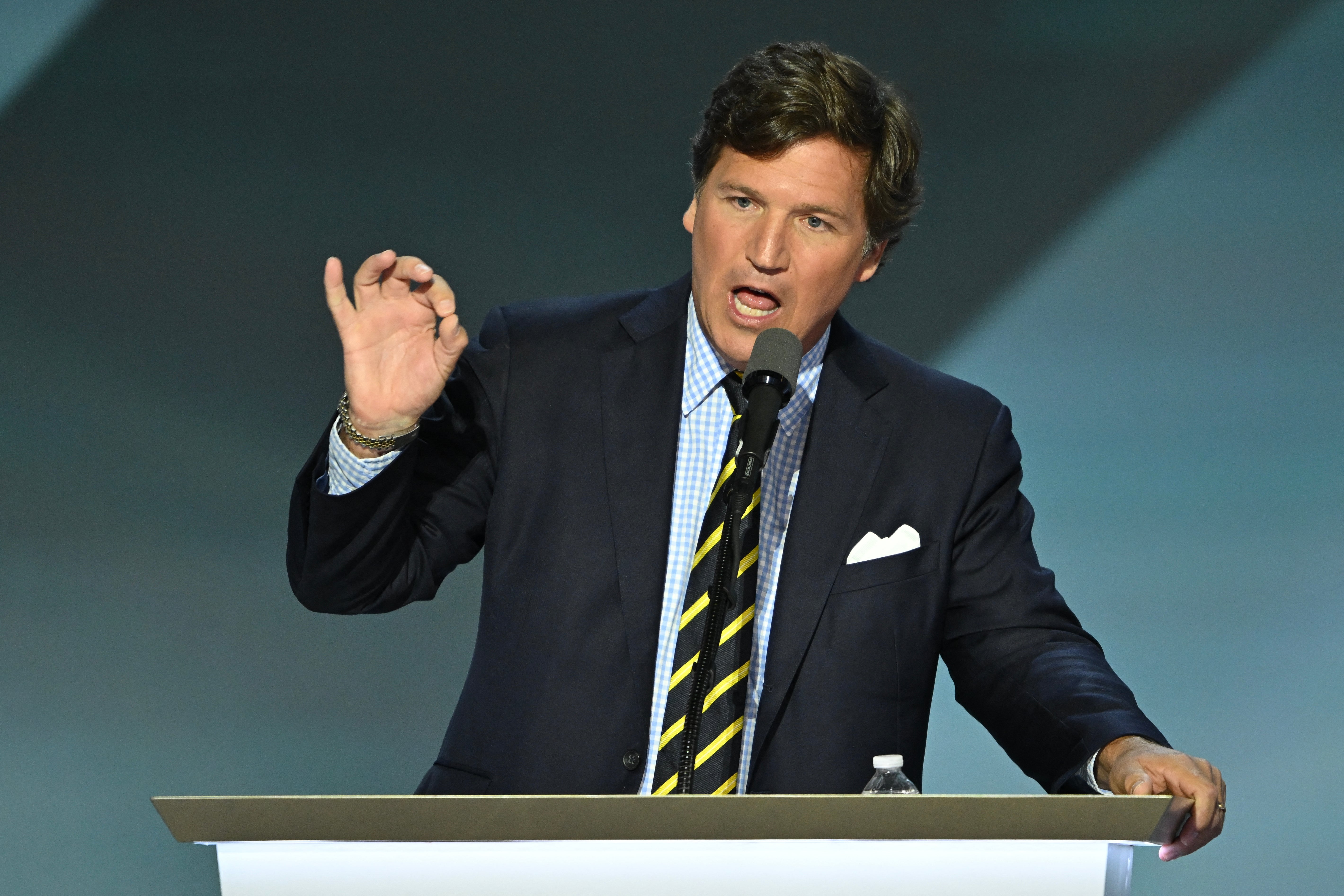 A idiosyncratic successful a suit speaks passionately astatine a podium during an event, gesturing with 1 manus raised and holding a microphone