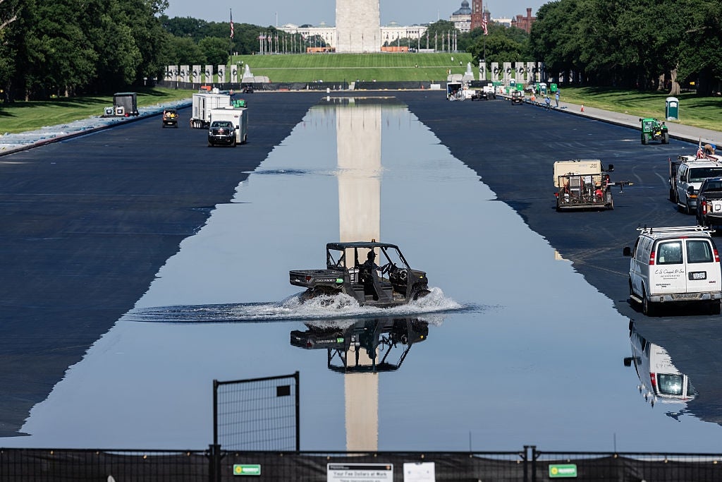 Kjøretøy som kjører gjennom reflekterende basseng nær Washington-monumentet, ser ut som på vann, med arbeidere og utstyr i bakgrunnen