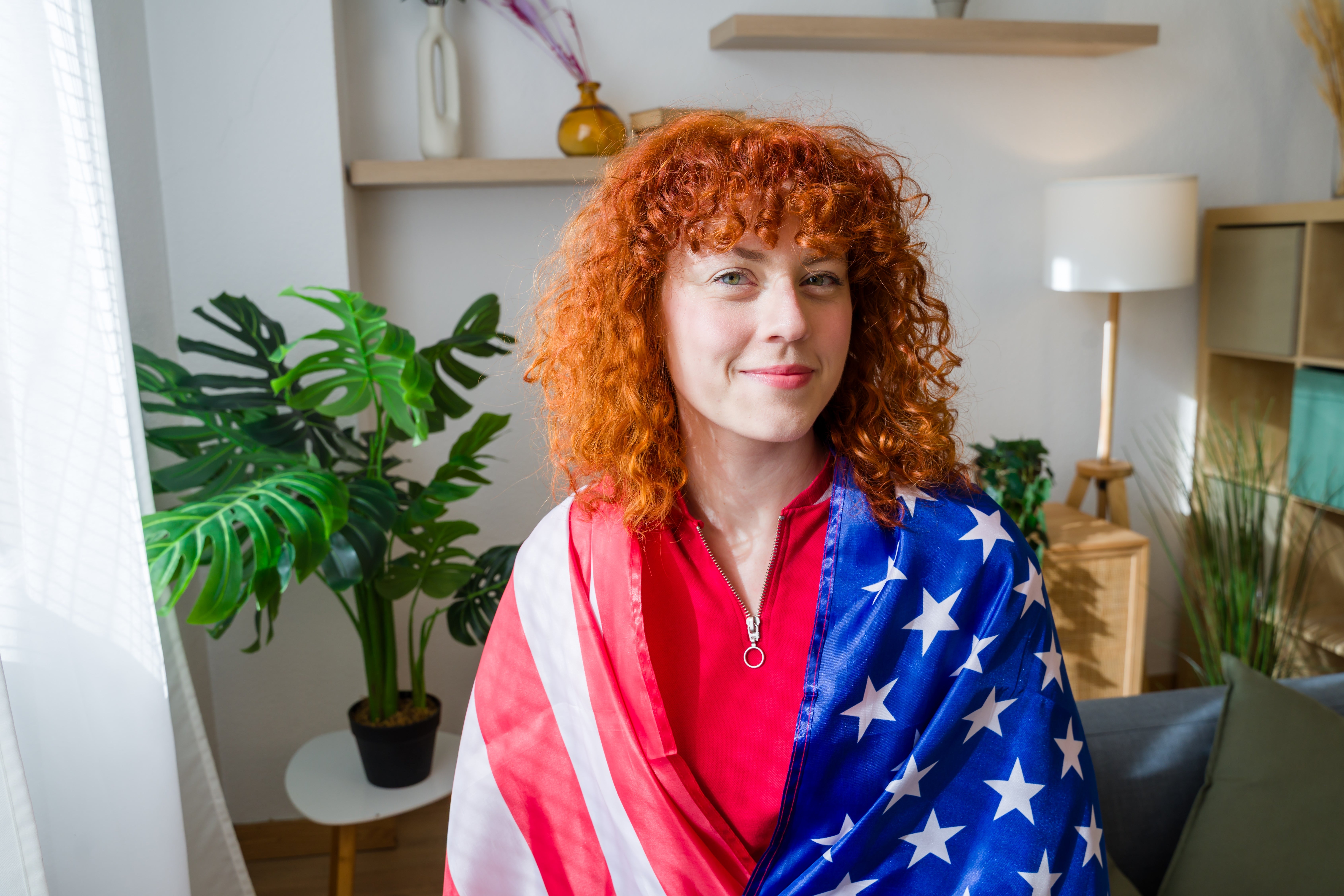 Person med krøllete hår drapert i stjernemønstret flagg, som sitter innendørs med planter, og formidler en selvsikker, patriotisk oppførsel