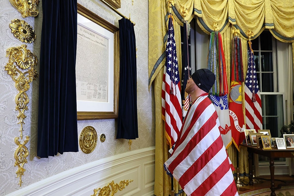 Person pakket inn i et amerikansk flagg stirrer på et historisk dokument i et utsmykket rom med flagg og innrammede bilder