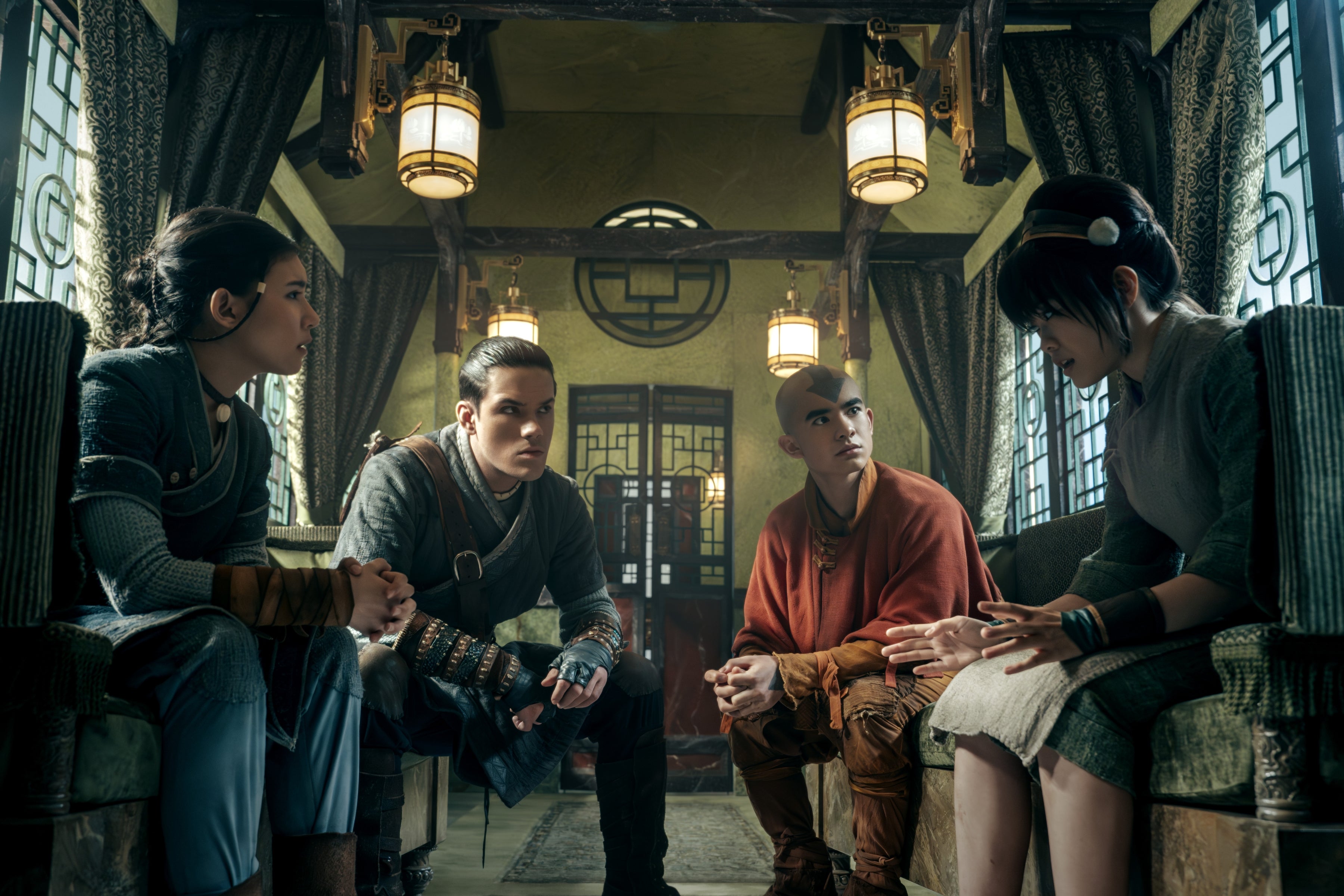 Four actors in detailed, warrior-style costumes sit in an ornate, traditional room, engaged in a serious conversation