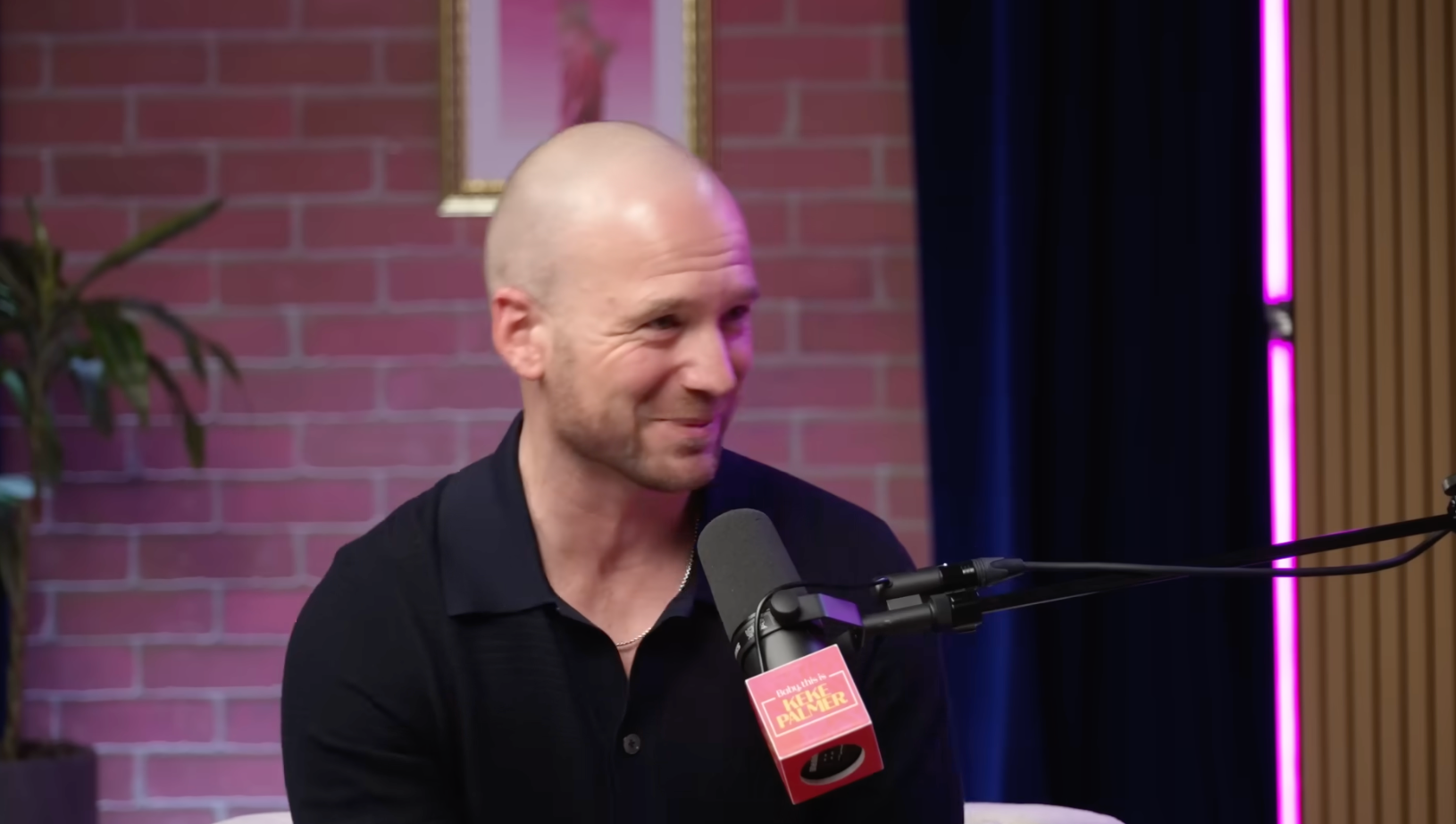 A idiosyncratic with a shaved caput is speaking into a podcast microphone, smiling. They are wearing a collared garment and seated indoors