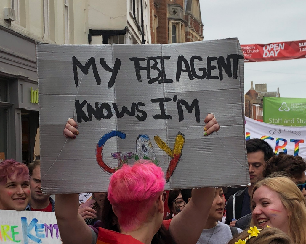 Person holding a handmade sign that reads, "MY FBI AGENT KNOWS I'M GAY," at a street event with people around