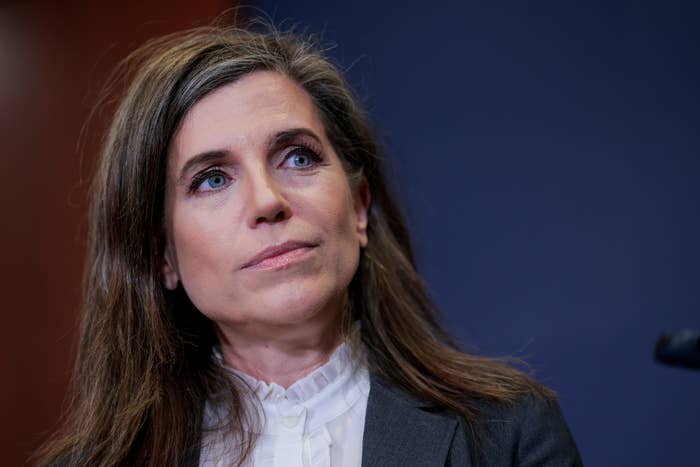 Person with agelong  hairsbreadth  wearing a suit   and ruffled blouse, looking thoughtfully to the broadside  against a plain backdrop