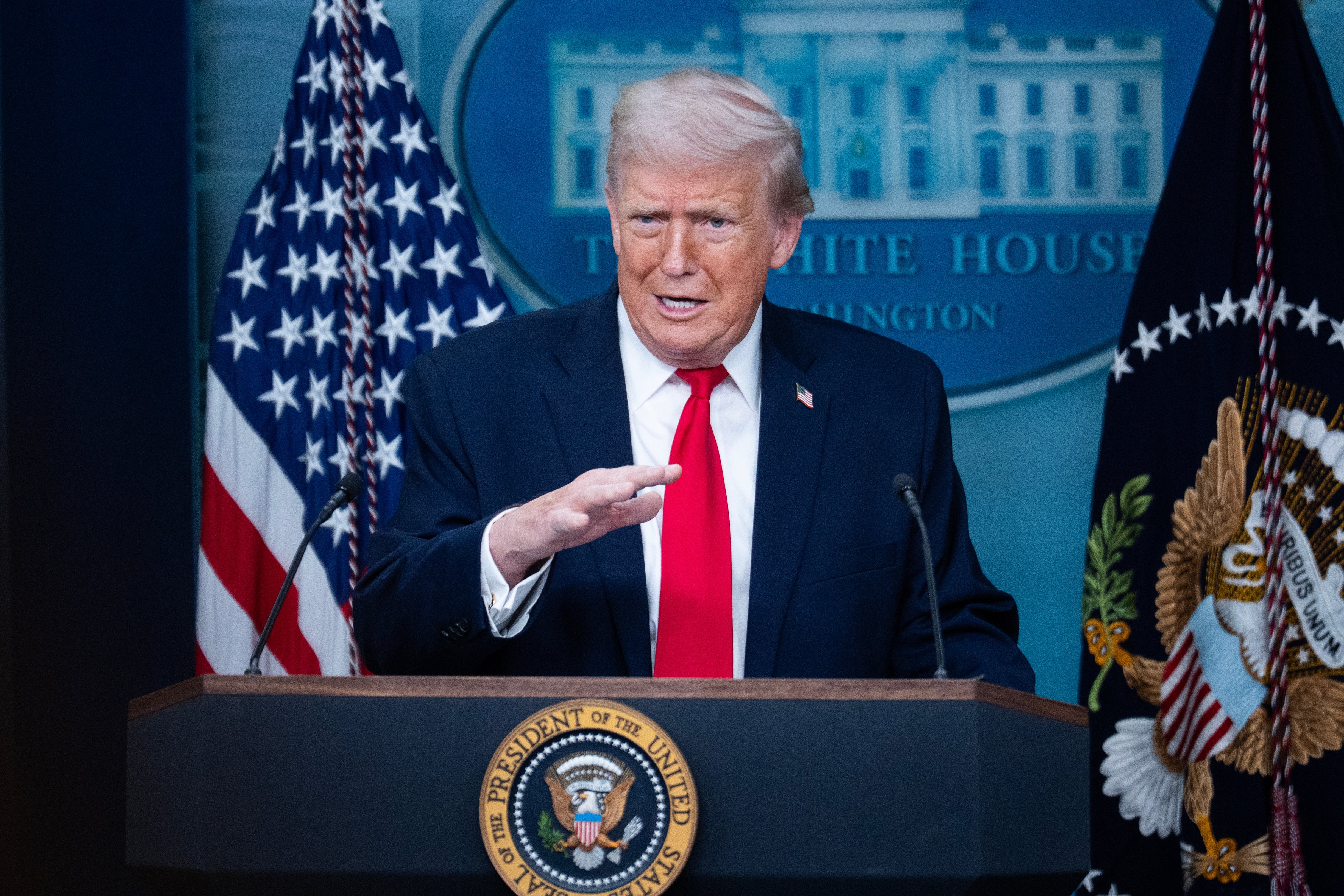 En person i dress og rødt slips snakker på et podium med presidentseglet, satt mot et bakteppe av amerikanske flagg og et bilde av en regjeringsbygning
