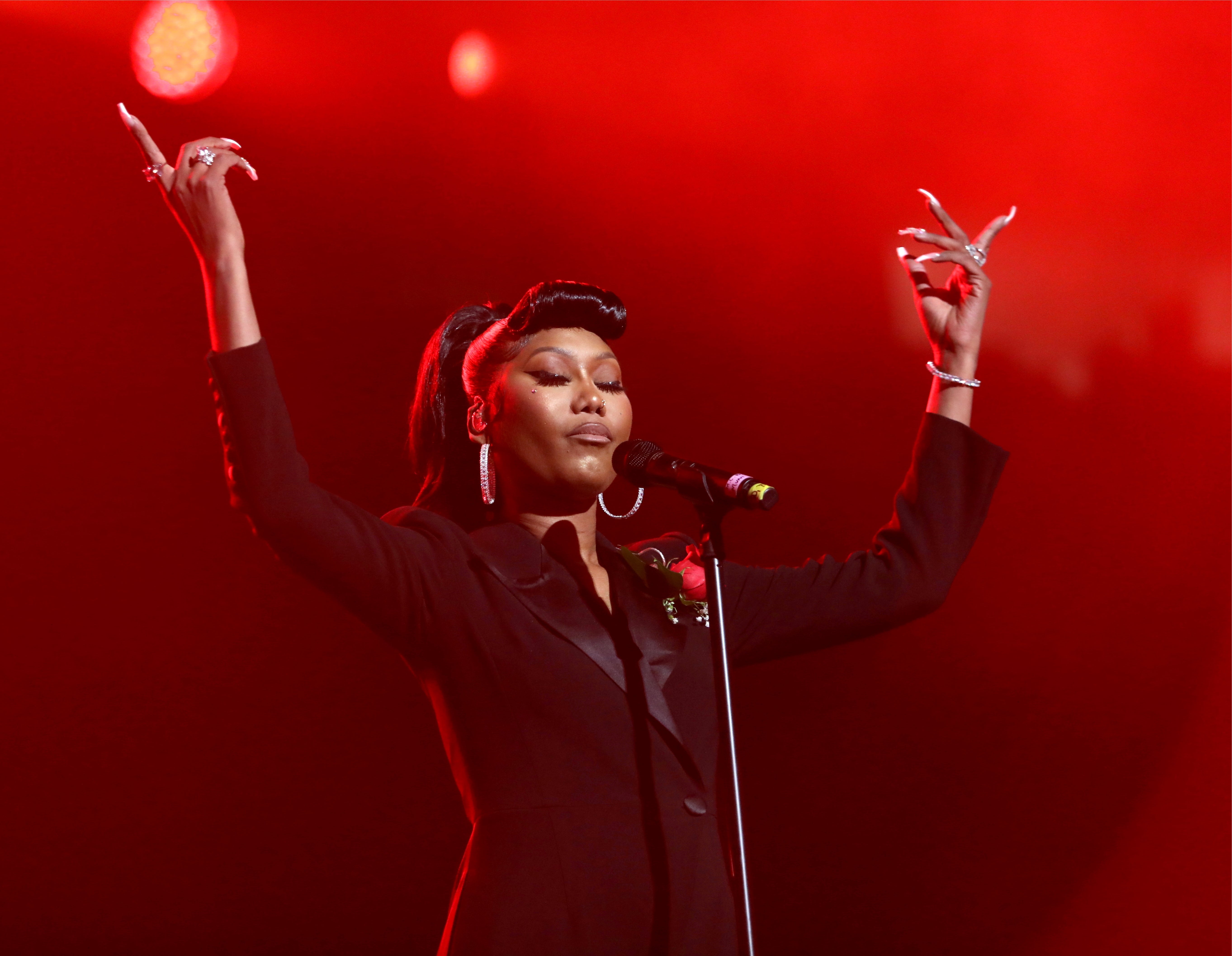 Celebrity performing connected signifier with an elegant achromatic outfit, agelong nails, and arms raised, nether melodramatic reddish lighting