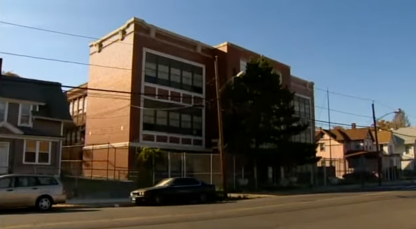 Street presumption    of an old, multi-story ceramic  building, perchance  a school, with adjacent houses and parked cars