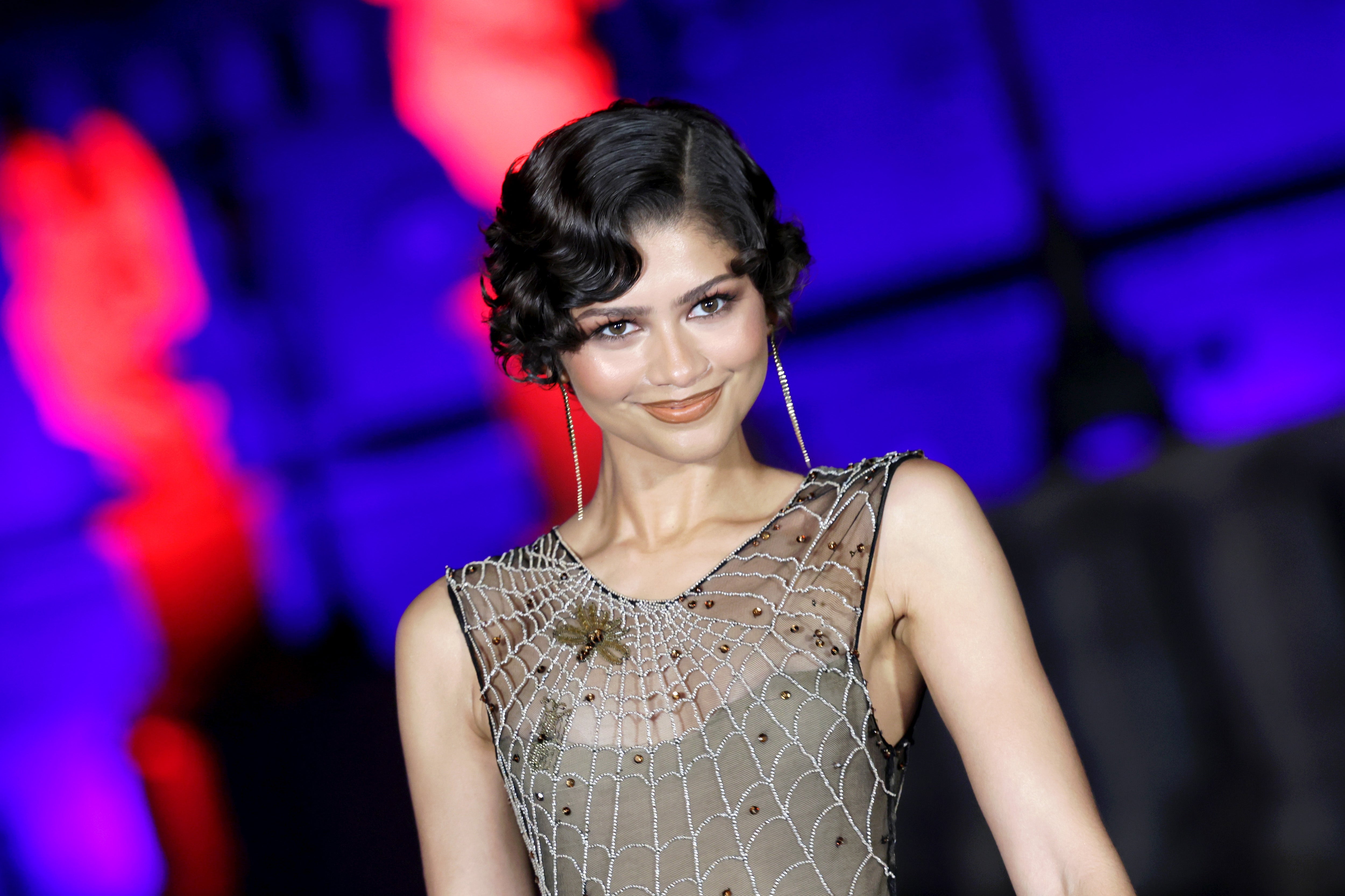 Zendaya i en elegant, nettmønstret ren kjole, smilende på en rød løper med elegante øredobber